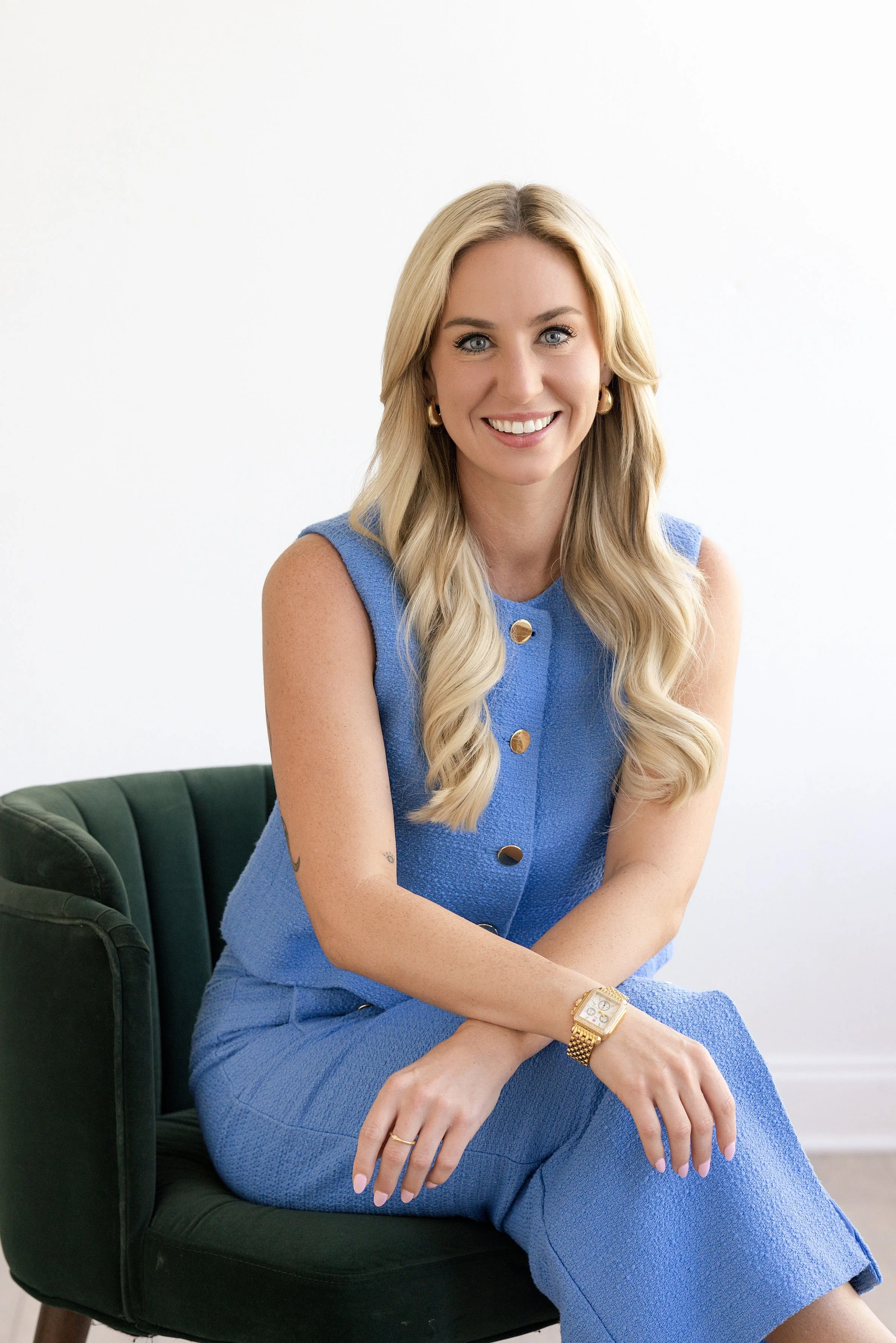 A woman with long blonde hair, wearing a blue sleeveless textured dress with gold buttons, sitting on a black chair with her arms crossed. She is smiling and wearing gold hoop earrings, a gold watch, and a gold ring. The background is plain white.