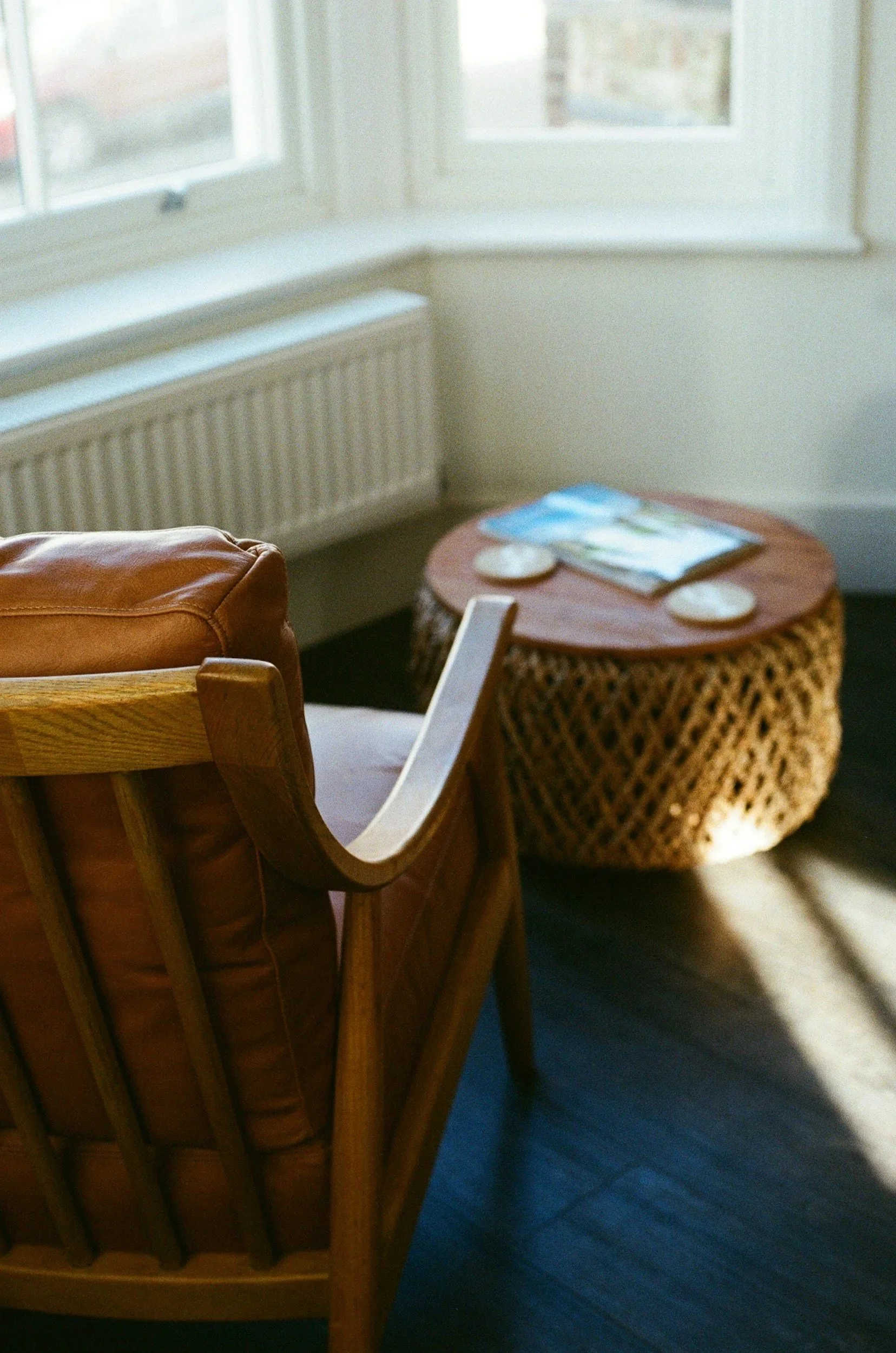 Close-up of a wooden chair with a leather cushion in a room with a window and a wicker side table with magazines and coasters.