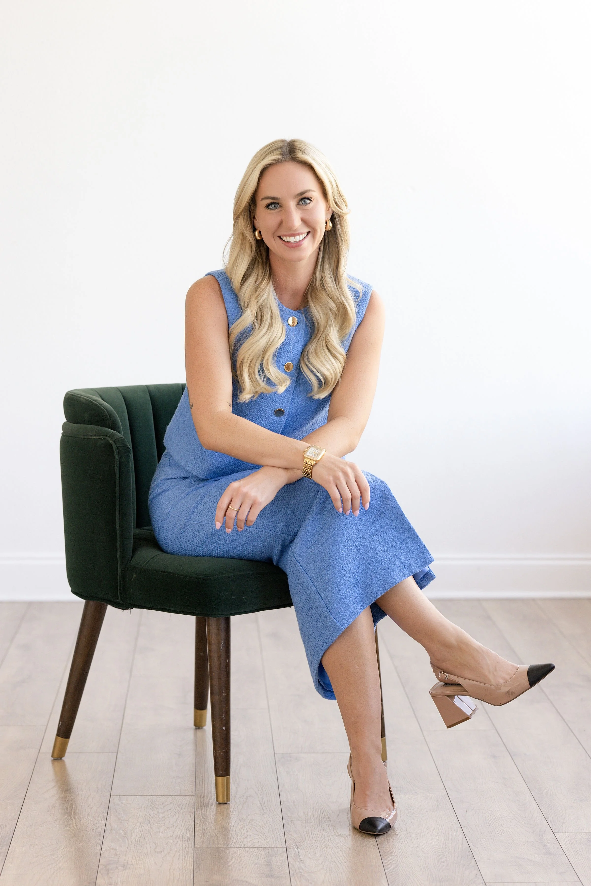 A woman with long blonde hair sitting on a green office chair with wooden legs, smiling at the camera, against a plain white background. She is wearing a sleeveless blue dress, beige high heels, and gold jewelry, including earrings, a bracelet, and a watch.