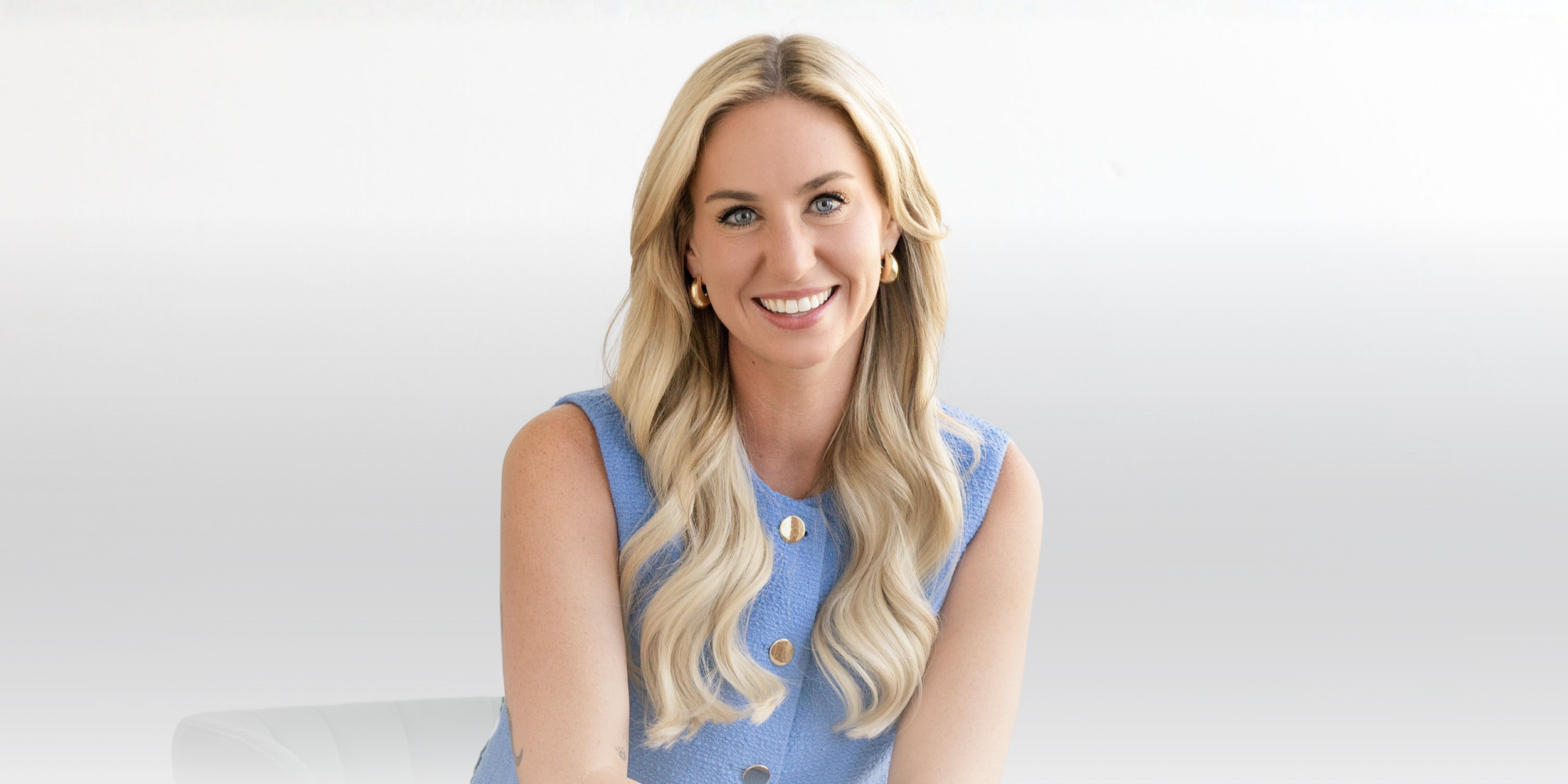 A woman with long blonde hair, wearing a sleeveless blue top with buttons, smiling at the camera against a plain white background.