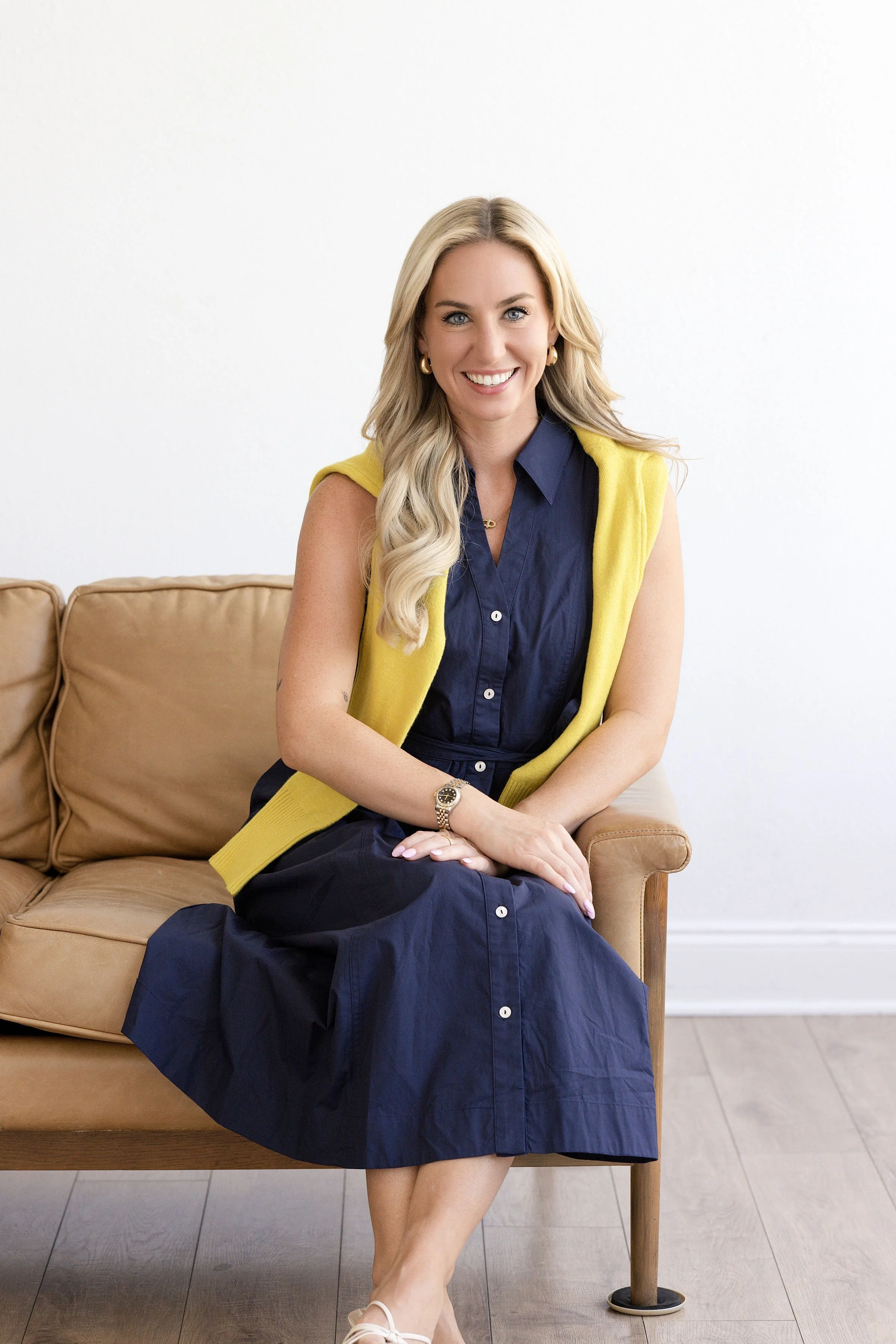 A woman with blonde hair smiling, wearing a navy blue dress and a yellow sleeveless cardigan, sitting on a tan sofa with her legs crossed, against a white wall background.