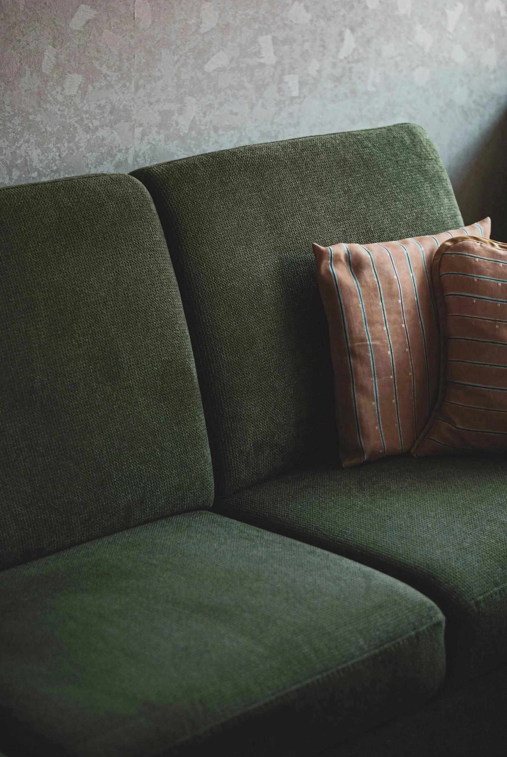 Close-up of a green upholstered sofa with two brown striped pillows resting against the backrest, set against a textured wall.
