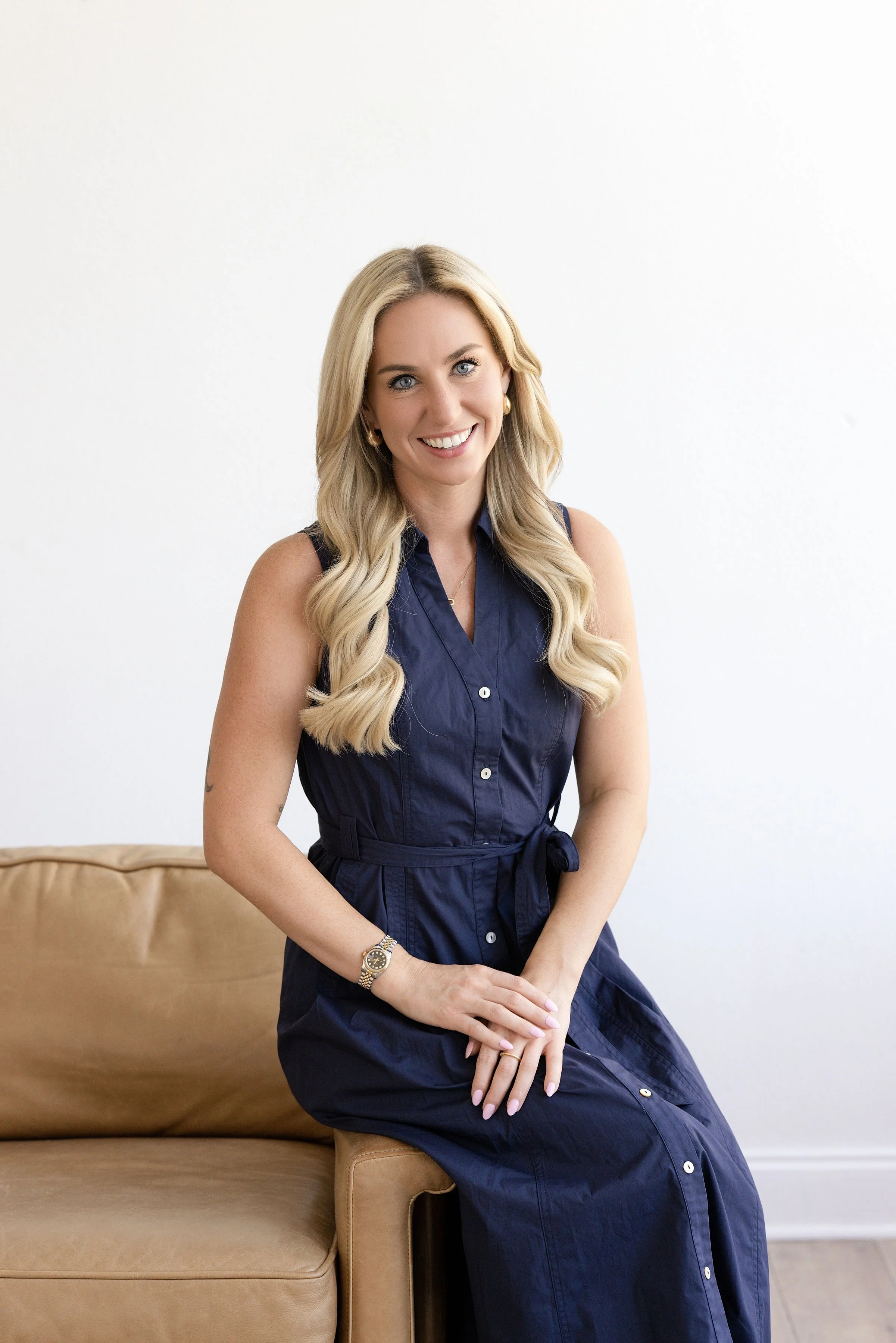 Savannah Goudreau, a woman with long blonde hair, wearing a sleeveless navy blue dress, sitting on a tan leather couch with her hands clasped on her lap, smiling at the camera in a bright room with a white wall.