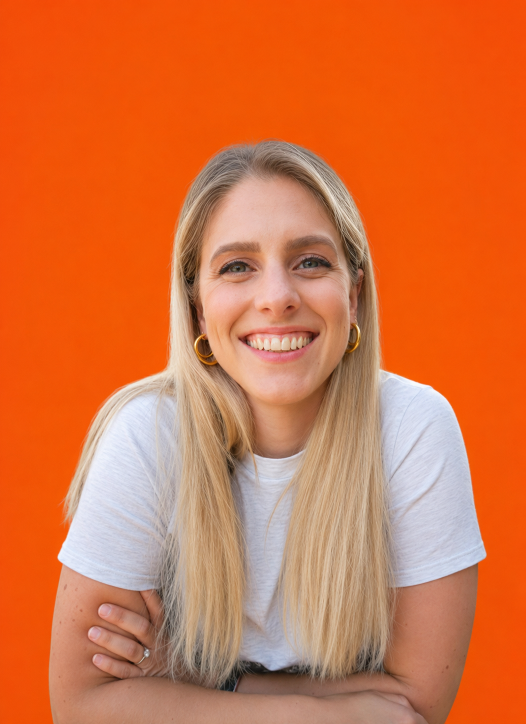 Smiling woman with long blonde hair, wearing gold hoop earrings and a white t-shirt, posing with crossed arms against a bright orange background.