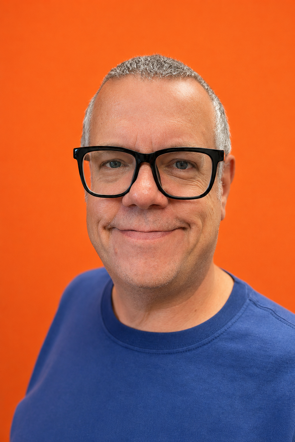 Smiling middle-aged man with short gray hair, wearing black-rimmed glasses and a blue shirt, against an orange background.