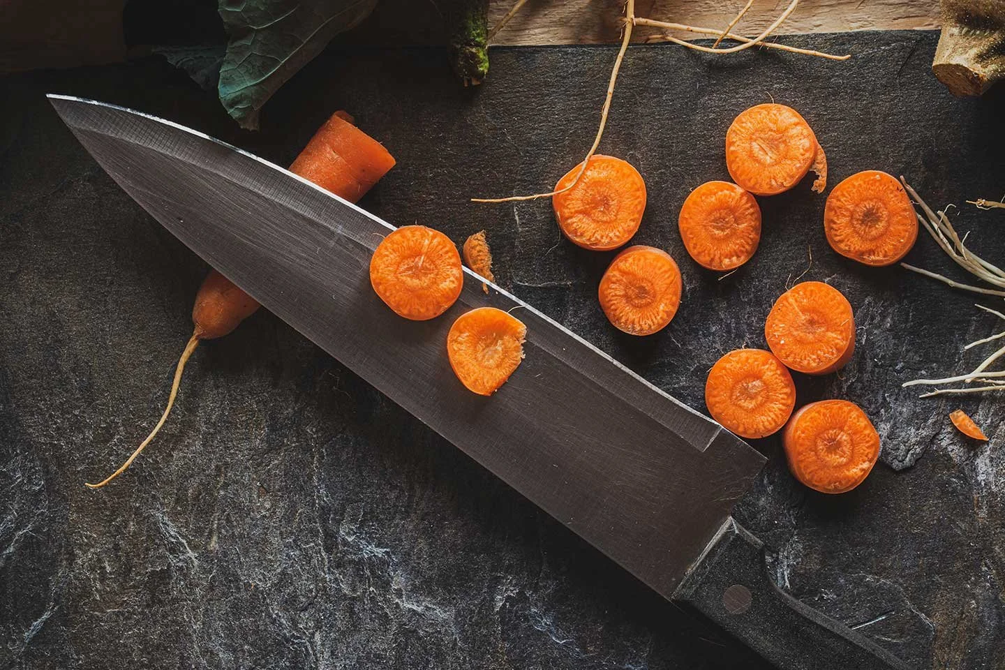 Fresh orange carrots, some sliced and others whole, with a cleaver on a dark textured surface. Dried twigs and green leaves are also visible.