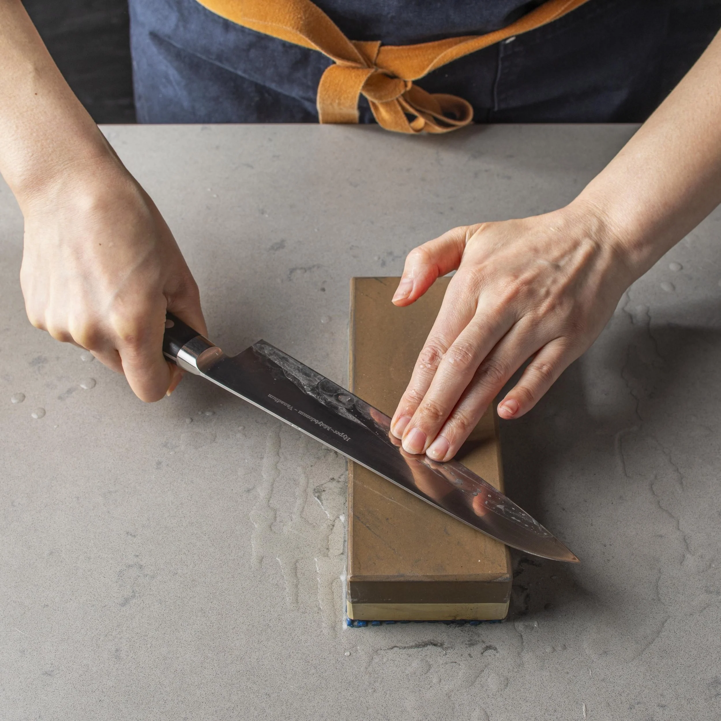 Person with light skin using a hammer to hit a chisel on a large rectangular stone or tile surface, with multiple square tiles or slabs in various shades of brown, gray, and dark brown arranged vertically.