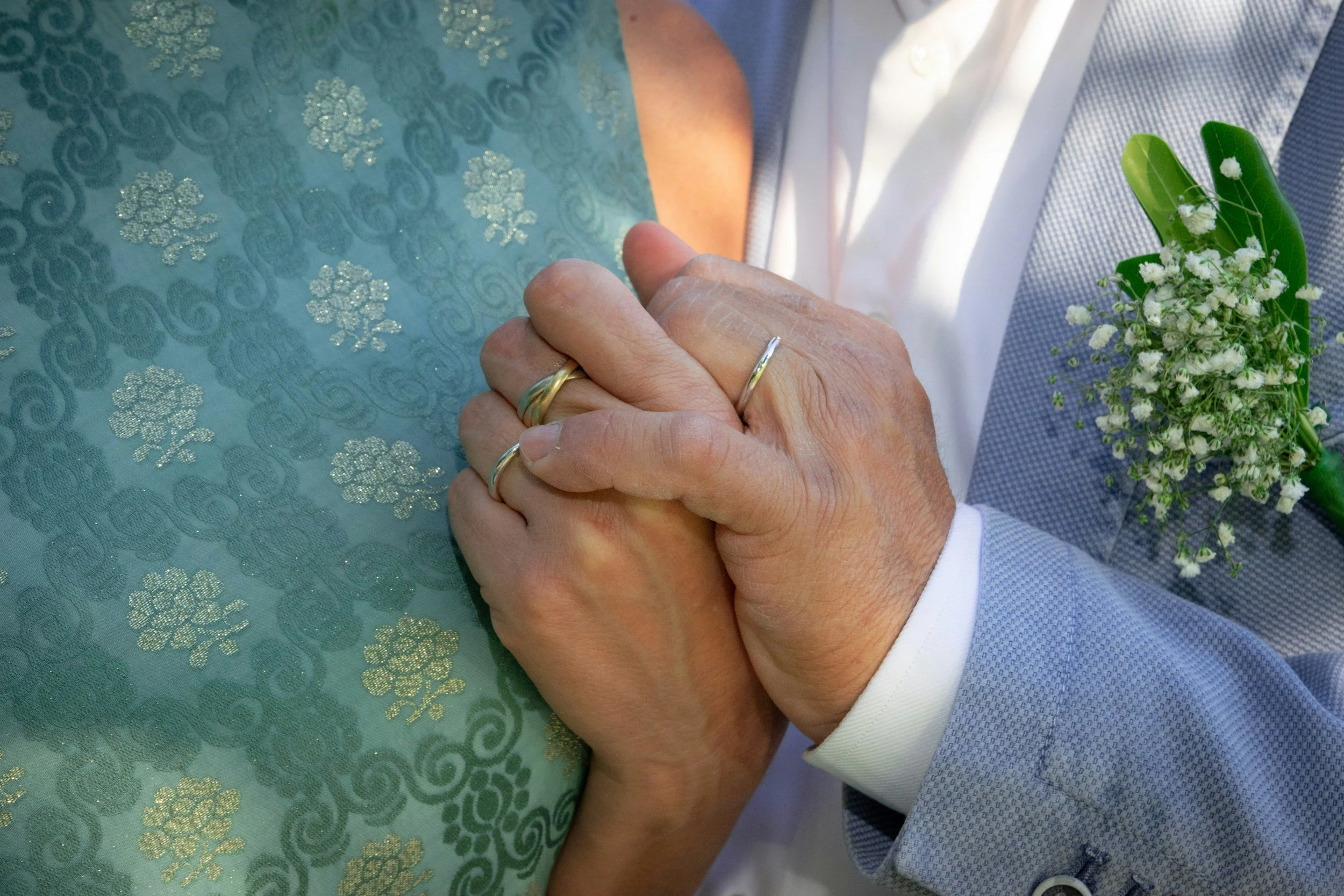 Close-up of an elderly couple holding hands during a wedding ceremony, with the bride wearing a light blue dress with silver floral embroidery and the groom in a light gray suit with a white shirt, holding a bouquet of white flowers.