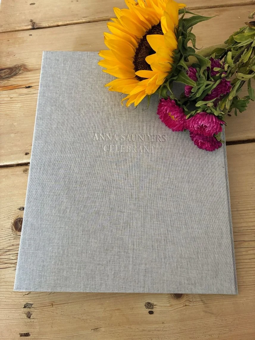 A gray hardcover book titled 'Anna Saunders Celebration' is placed on a wooden surface. A bouquet consisting of a sunflower with yellow petals and dark center, along with pink flowers, is resting on the top right corner of the book.