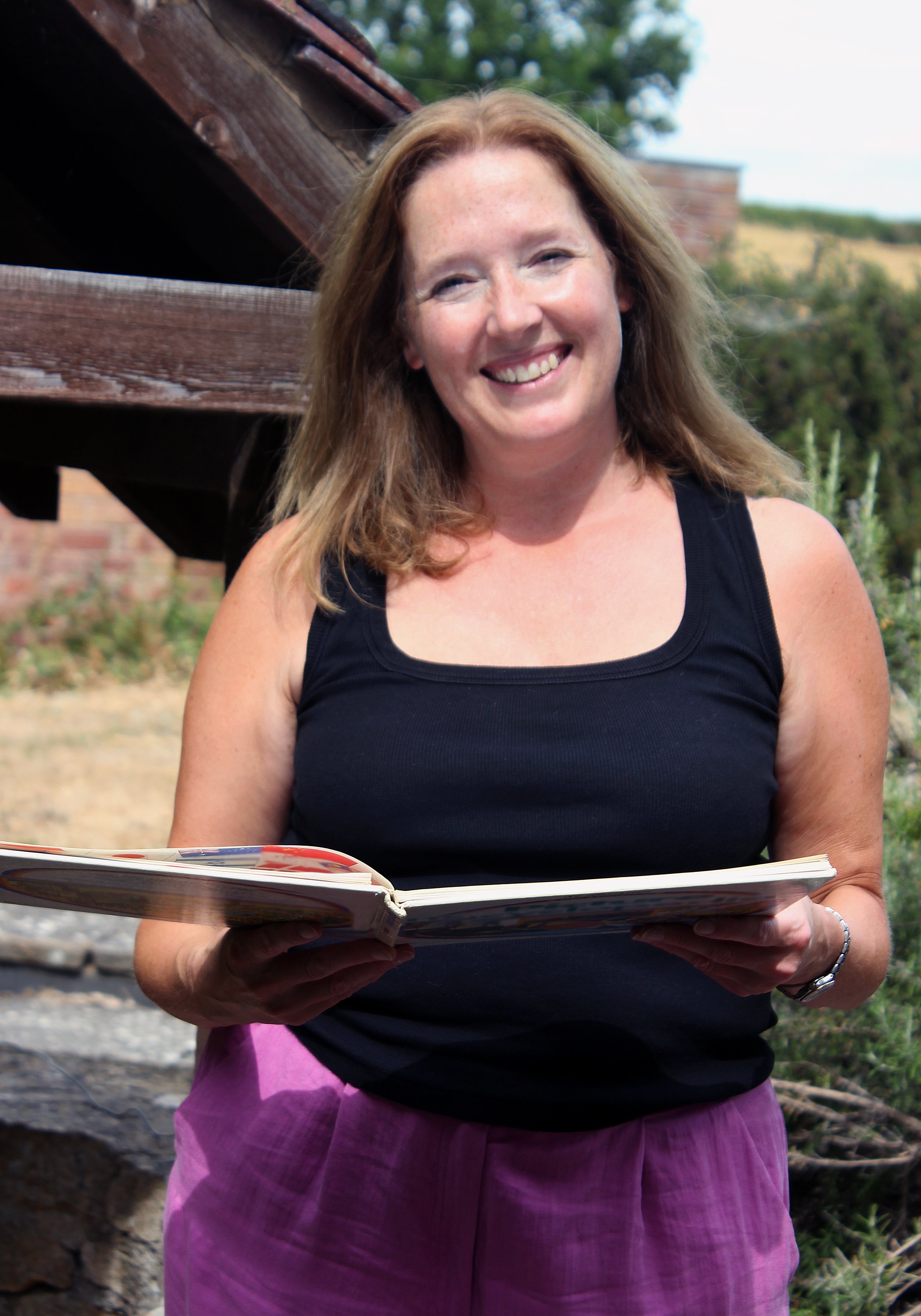 Anna, wearing a black tank top and pink trousers, smiling while holding an open folder outdoors.