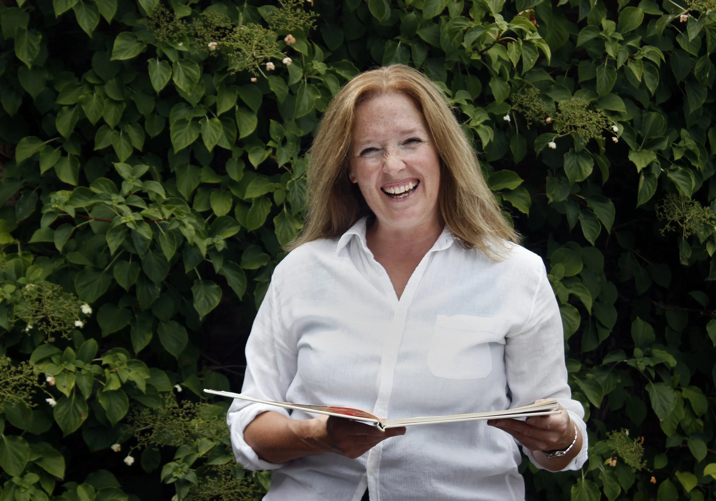 Anna, smiling, wearing a white shirt, holding a folder, standing outdoors in front of green leafy bushes.