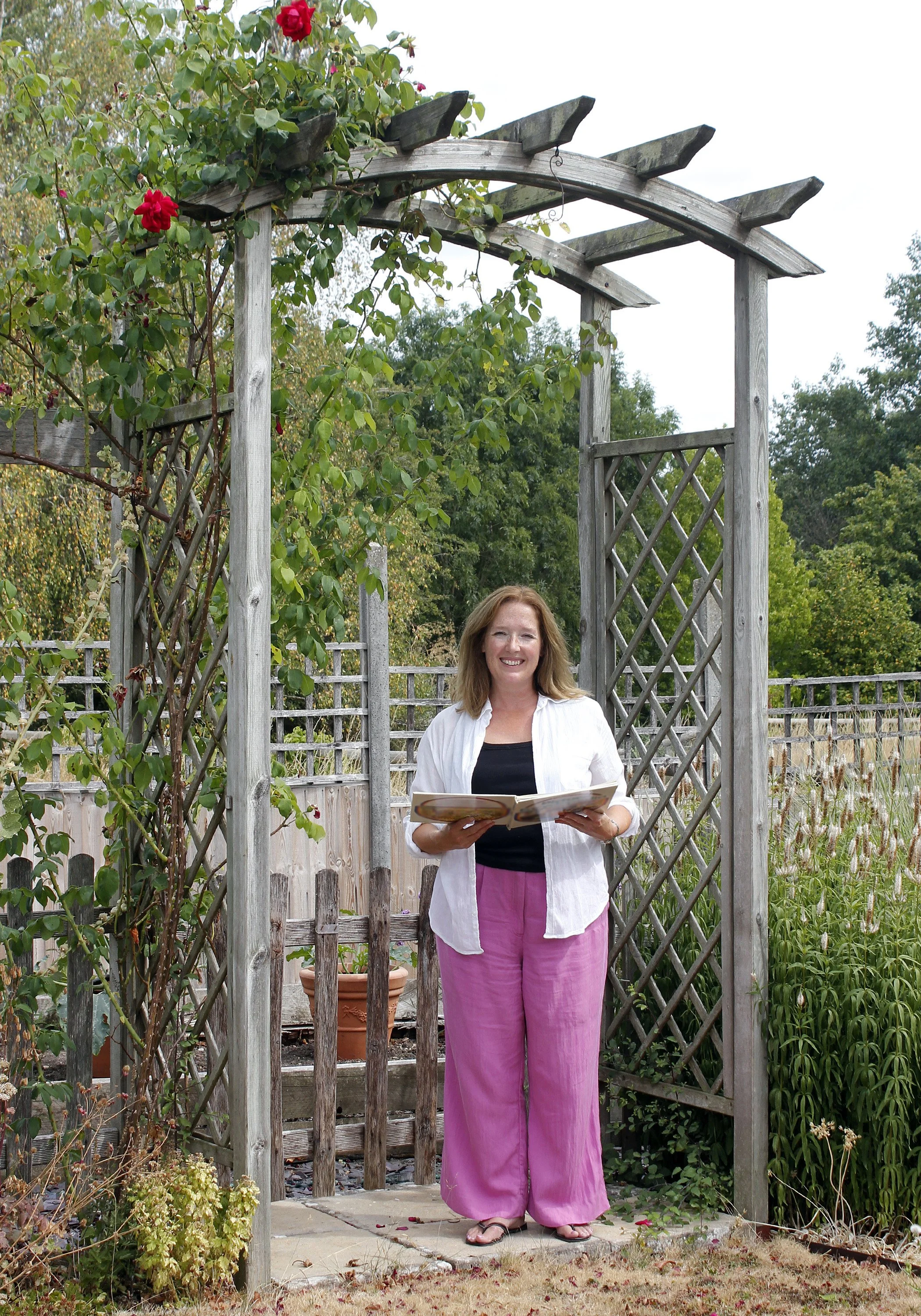 Anna, standing in a garden under a wooden archway, holding a book, wearing a white shirt, pink pants, and sandals, with green plants and a fence in the background.