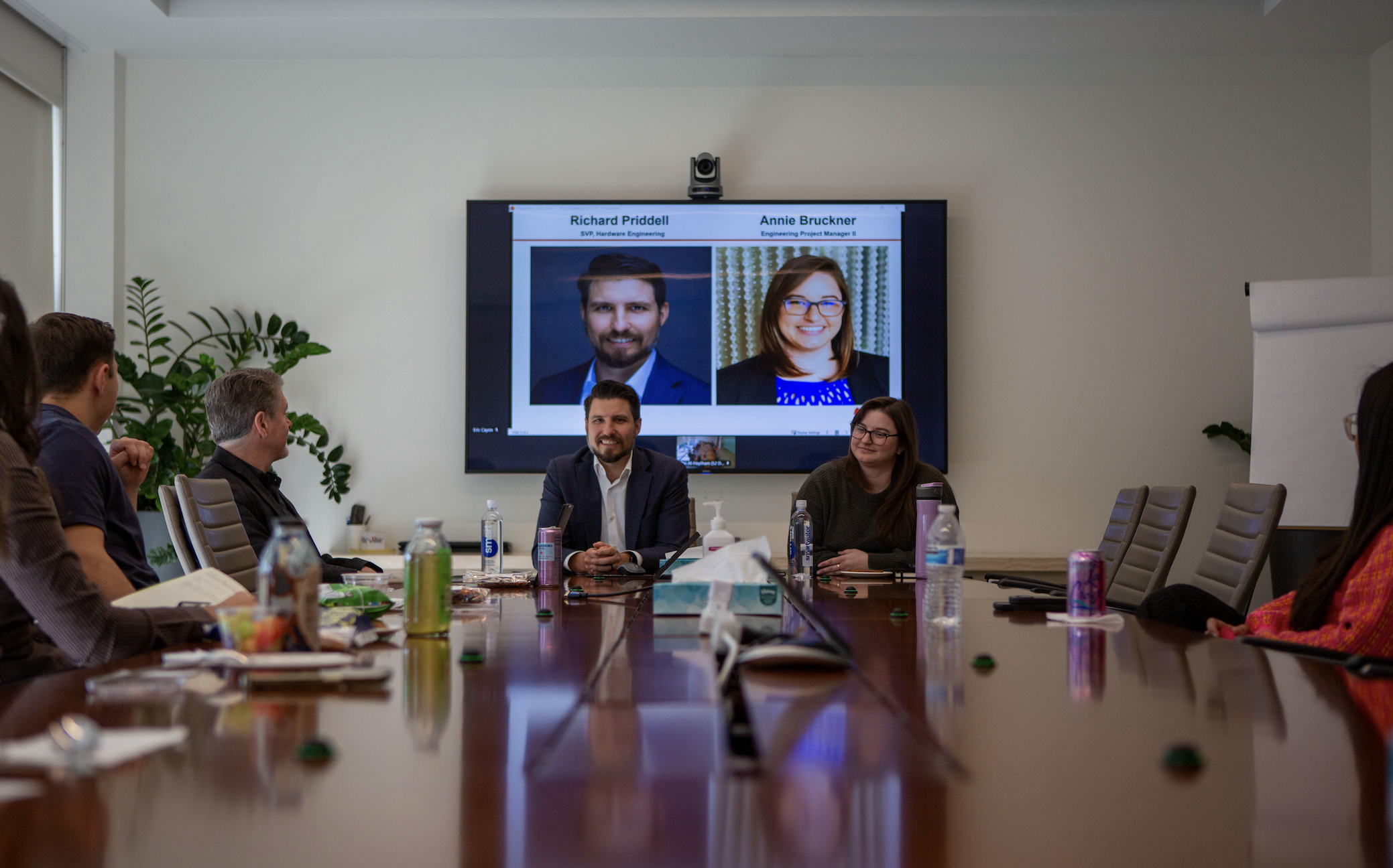 Business meeting in a conference room with people seated around a long table. A large screen at the front displays two headshots of a man with a beard and a woman with glasses, along with their names and titles.