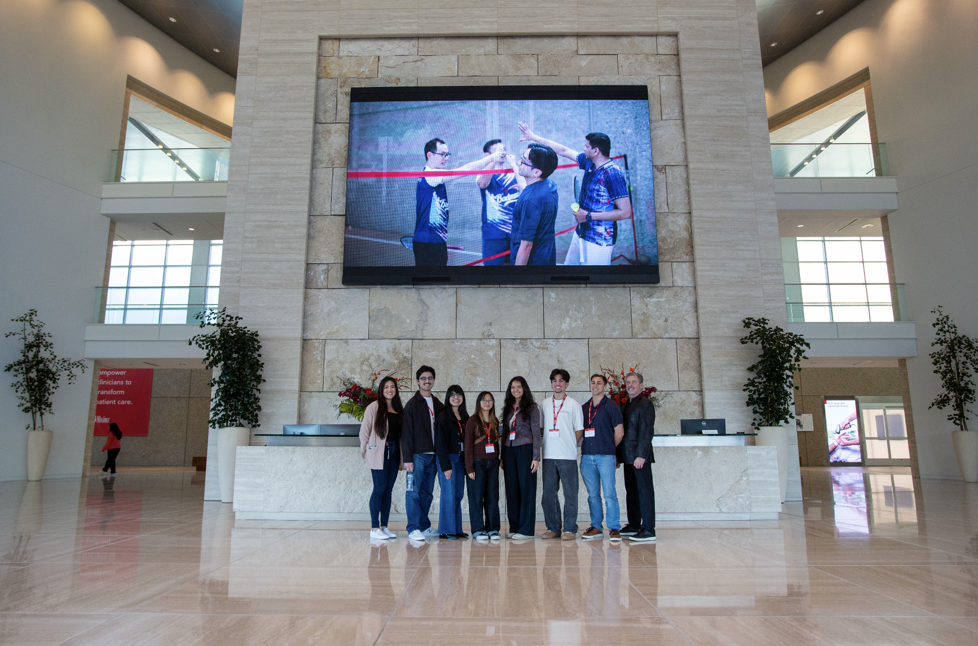Group of eight young adults standing together indoors in front of a large stone wall with a big digital screen showing four young men playing badminton. The group is smiling for the photo. There are plants and a reception desk in the background.