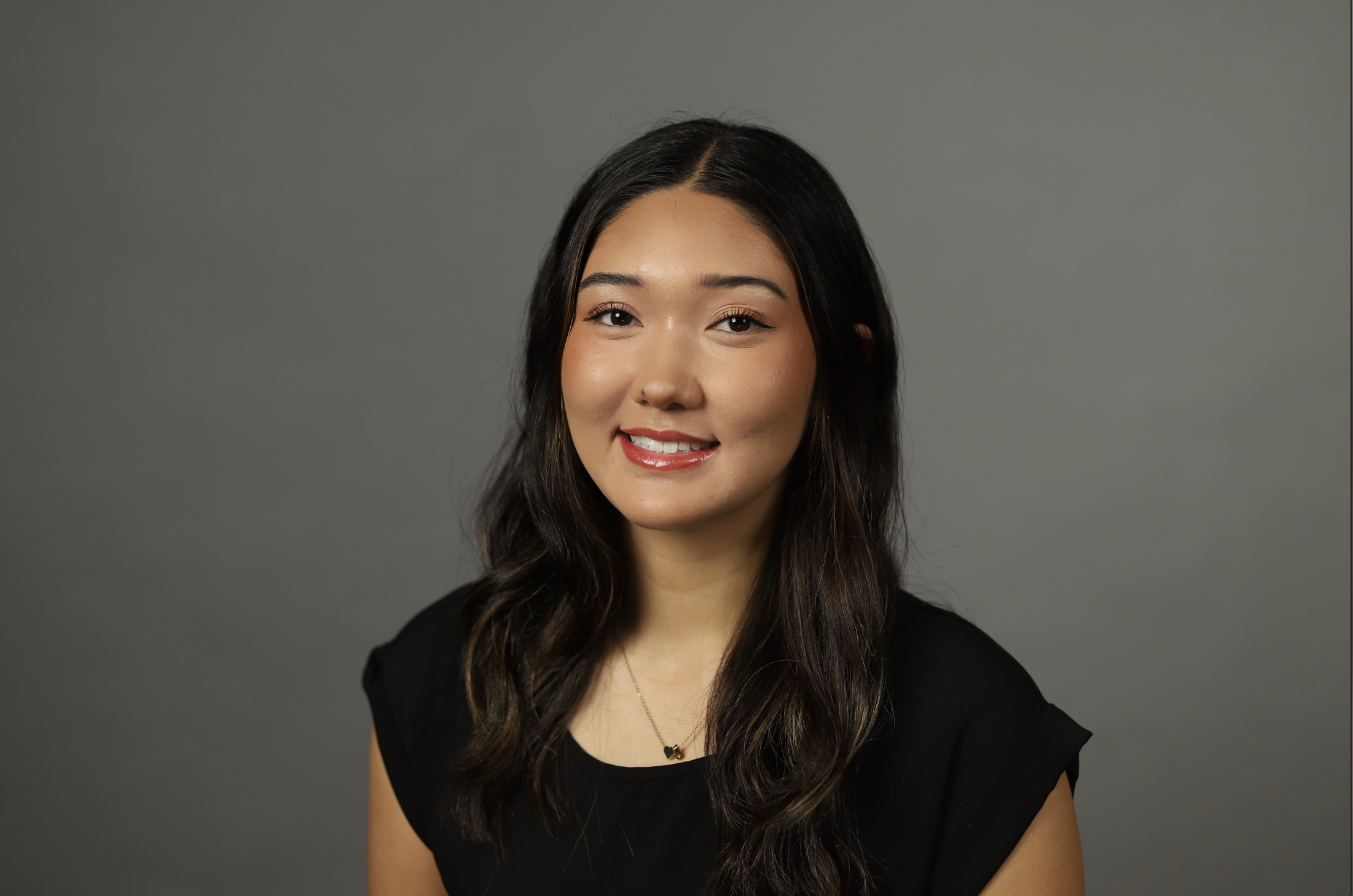 Portrait of a young woman with long dark hair, smiling, wearing a black top and a necklace, against a plain gray background.