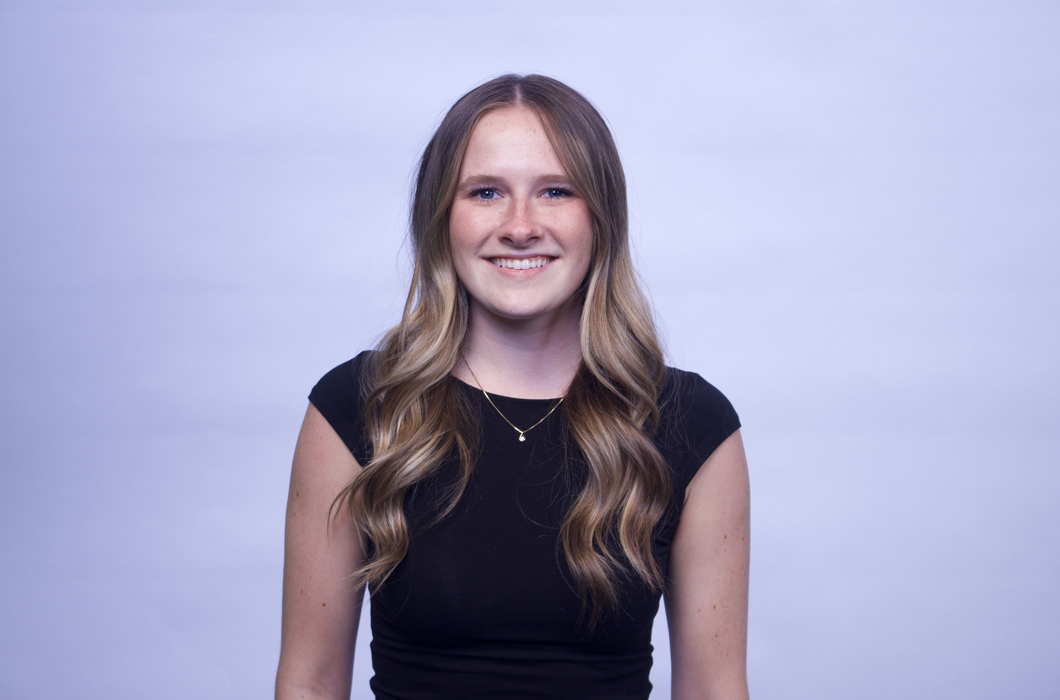 Portrait of a young woman with long, wavy blonde hair, blue eyes, wearing a black top and a gold necklace, smiling against a plain light blue background.