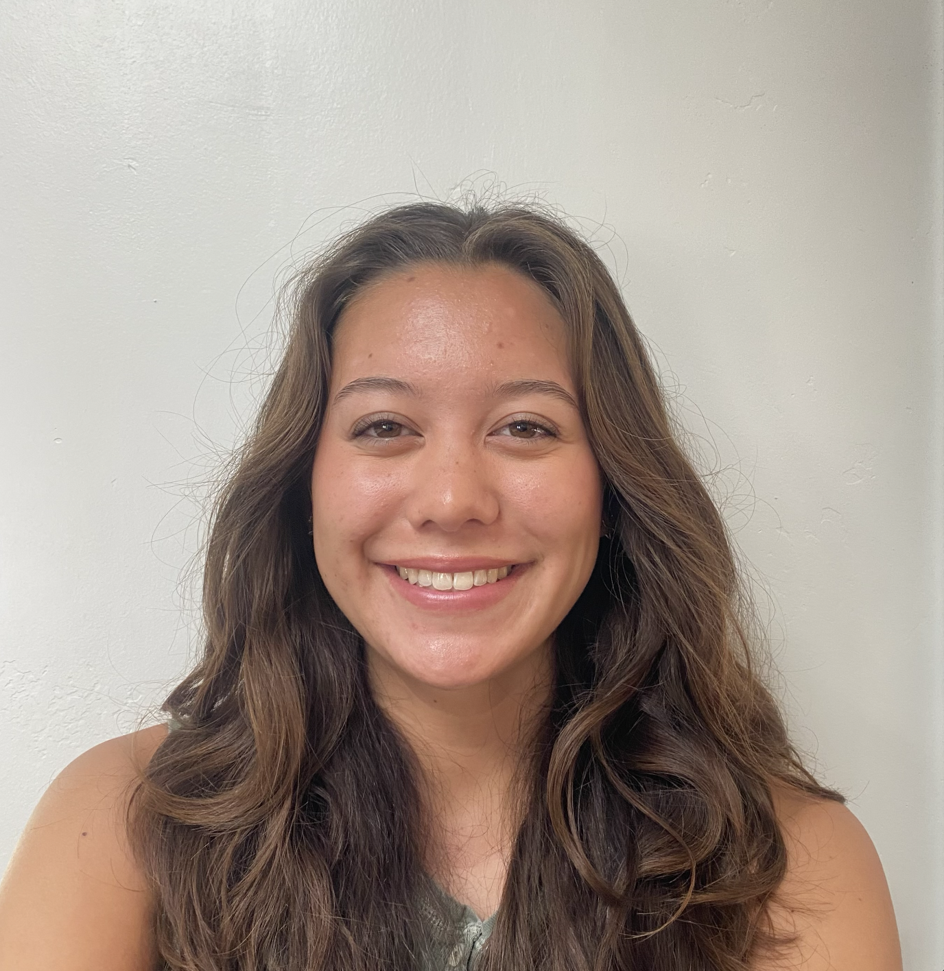 A young woman with long wavy brown hair smiling against a plain white wall.