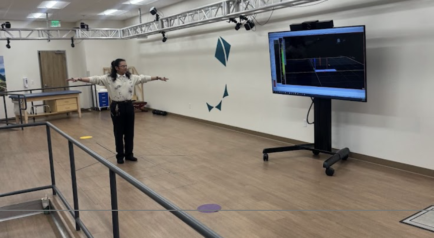A woman in a white sweater and black pants standing on a wooden floor with her arms outstretched in front of her, in a room with a large screen on a stand displaying a 3D modeling or scanning software. The room appears to be a testing or demonstration space with various equipment and a metal railing in the foreground.