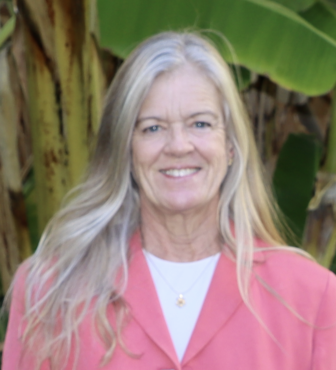 A smiling woman with long gray hair wearing a pink jacket and a white top, standing outdoors in front of green foliage.