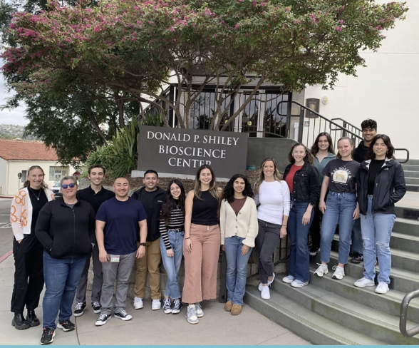 Group of diverse young people standing outdoors in front of the Donald P. Shiley Bioscience Center sign, with stairs and trees in the background.