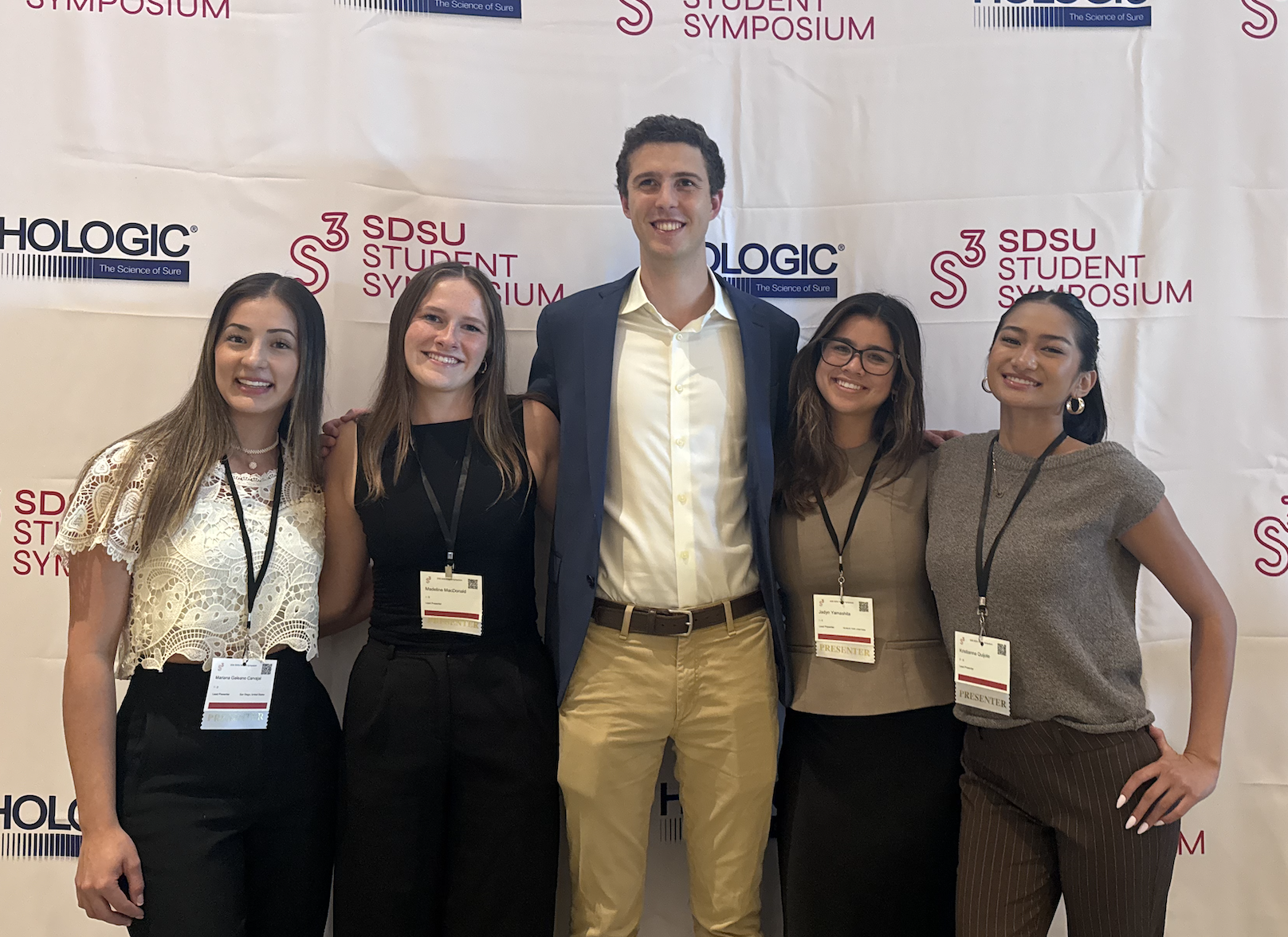 Group of five diverse young adults, four women and one man, standing together at a university event in front of a banner with logos for SDSU Student Symposium and Hologic. The women are wearing name badges, and the man is in a blazer and dress shirt.