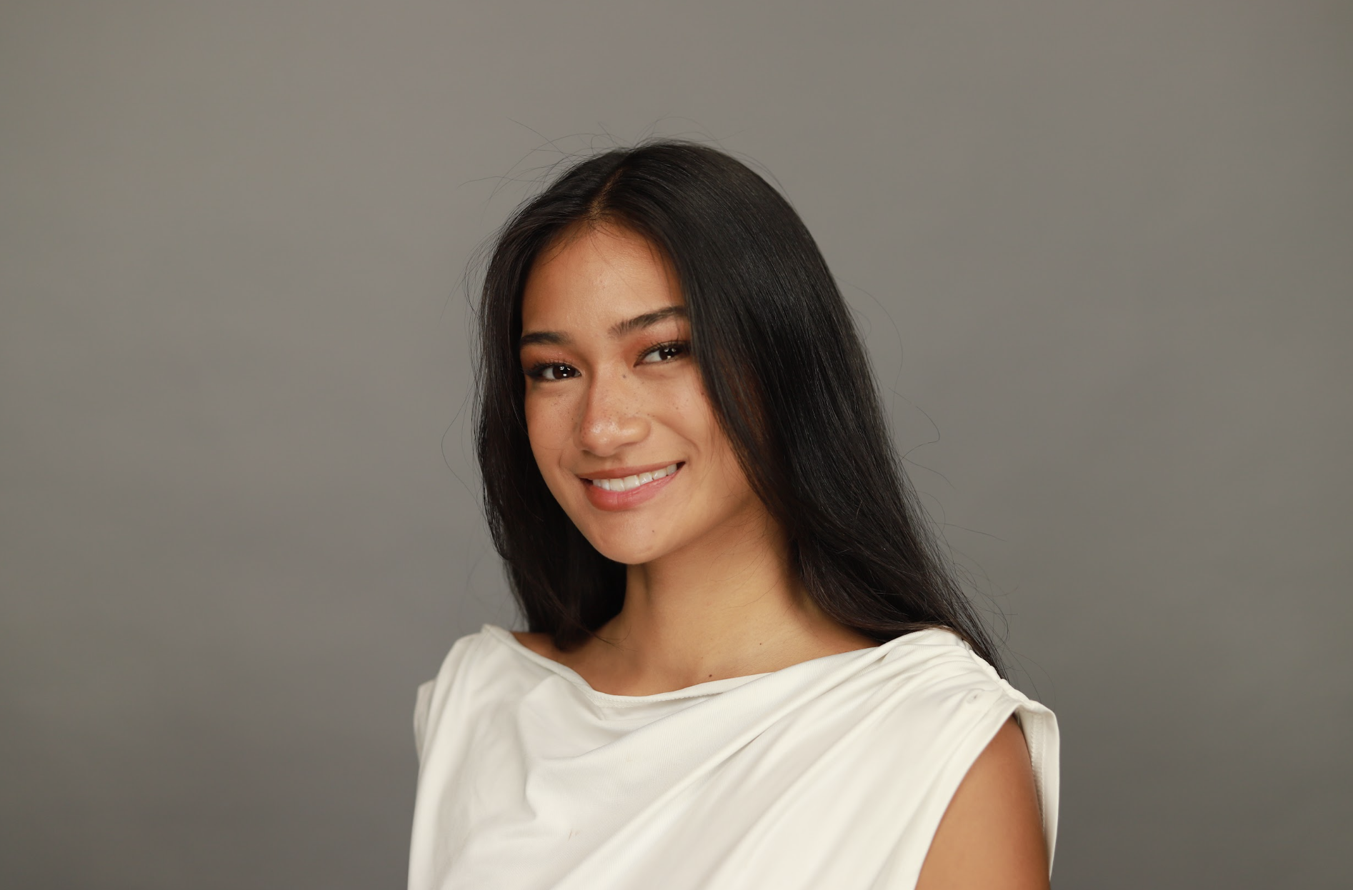 Portrait of a smiling woman with long dark hair, wearing a white top, against a plain gray background.