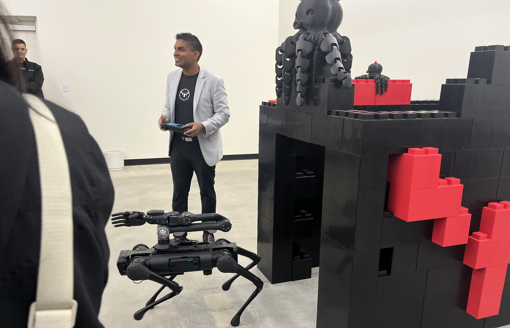 A man in a blazer and t-shirt giving a presentation to a group, with a large black and red LEGO structure and a robot arm in the foreground.