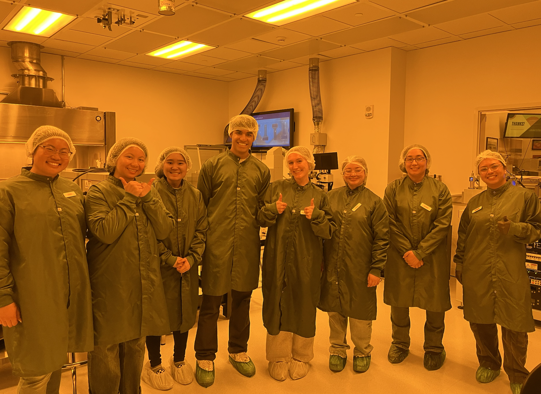 Group of eight people in a laboratory, all wearing green lab coats, hairnets, and shoe covers, smiling for the photo.