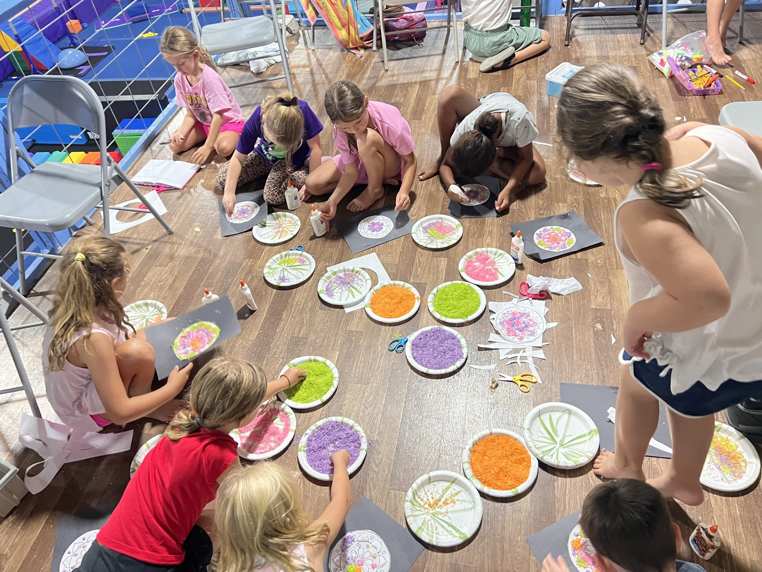 Children gathered around a table with paper plates, creating colorful art with glue, glitter, and markers in an indoor setting, possibly a classroom or craft area.