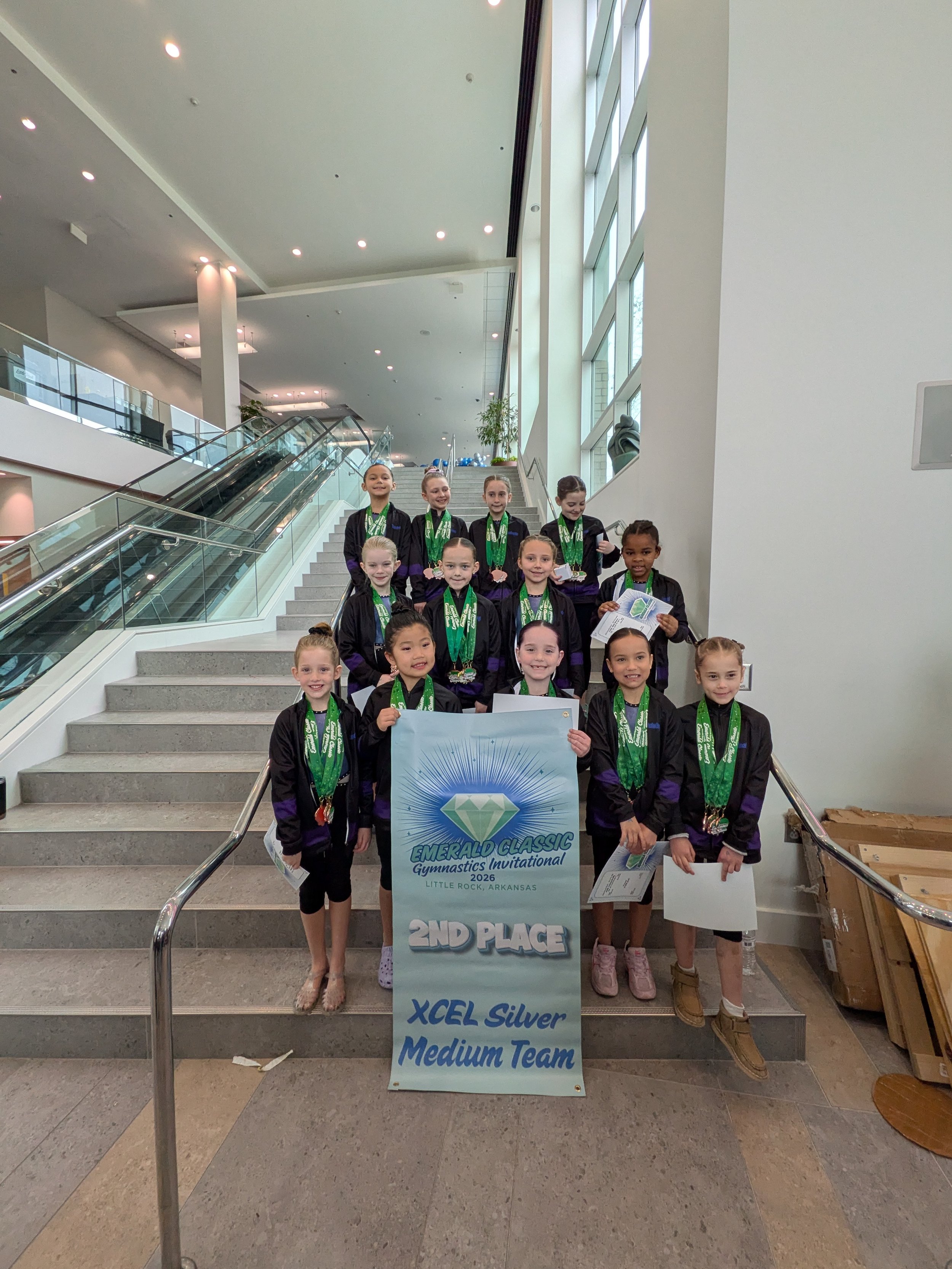 A group of young gymnasts holding medals and certificates, posing on a stairway inside a building with large windows. They are celebrating their second place win at the Emerald Classic Gymnastics Invitational 2026 in Little Rock, Arkansas. The sign in front indicates they are a medium team, XCEL Silver category.