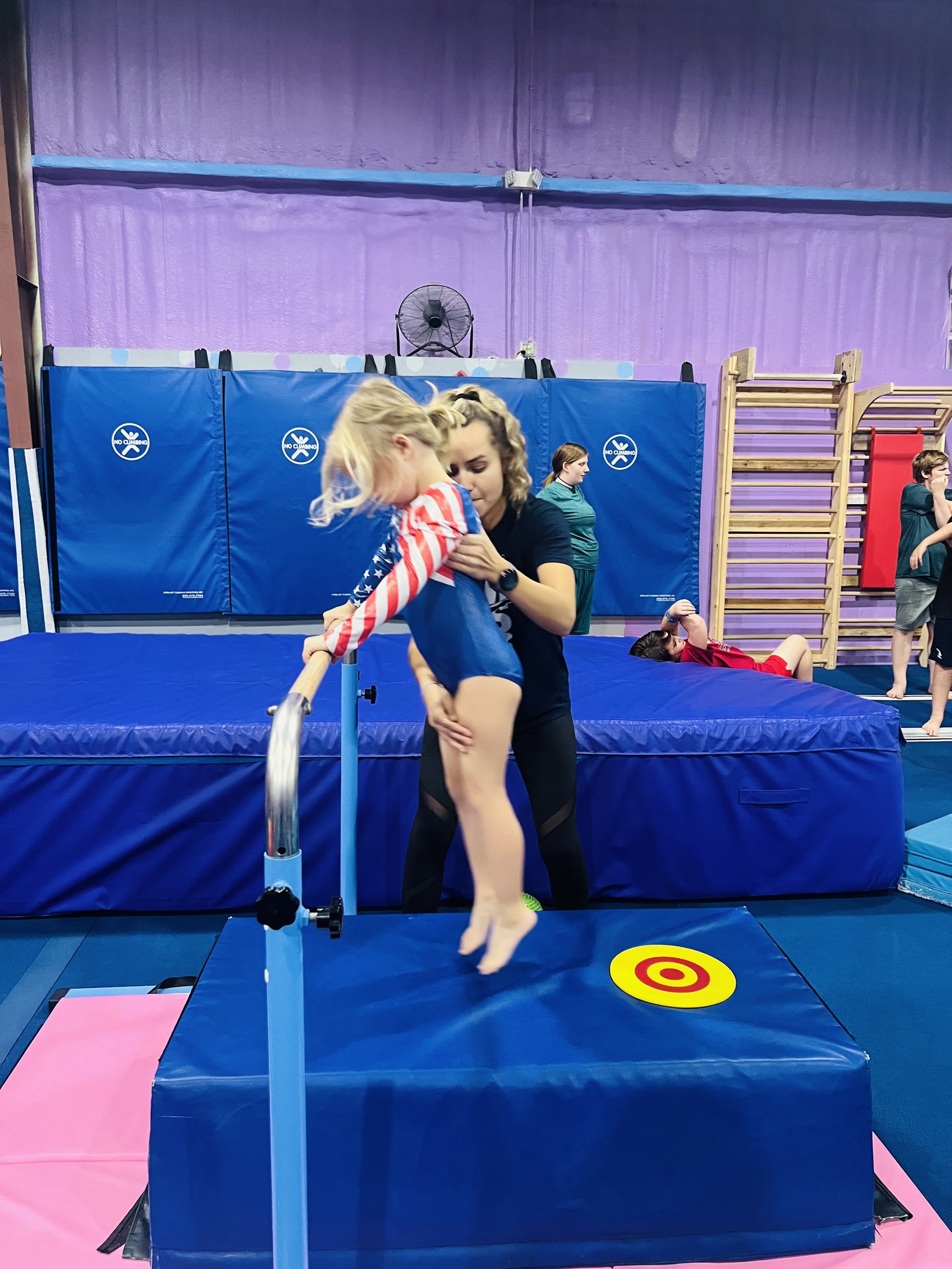 A young girl practicing gymnastics on a balance beam in a gym, with an instructor helping her. The gym has blue mats, purple walls, and wooden gym equipment. There is a target on the floor and a safety trampoline underneath the beam.