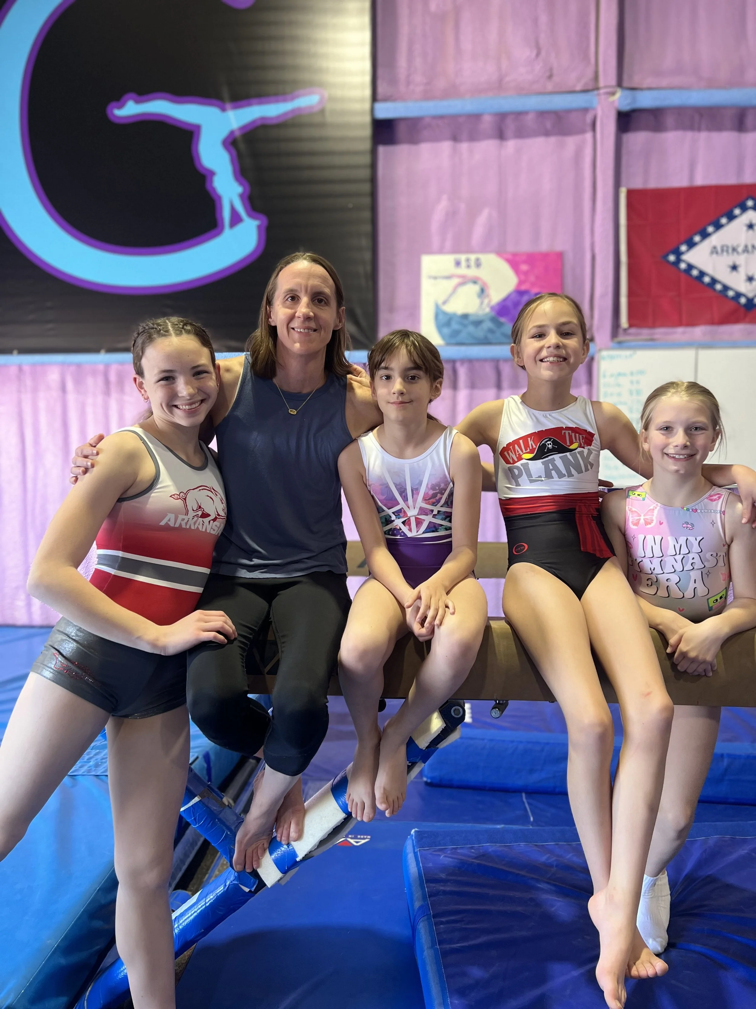 A group of five people, including four young girls and an adult woman, sitting together in an indoor gymnastics or trampoline park with purple and pink wall decorations and banners in the background. The girls are wearing sports outfits and the adult woman is in a sleeveless top.