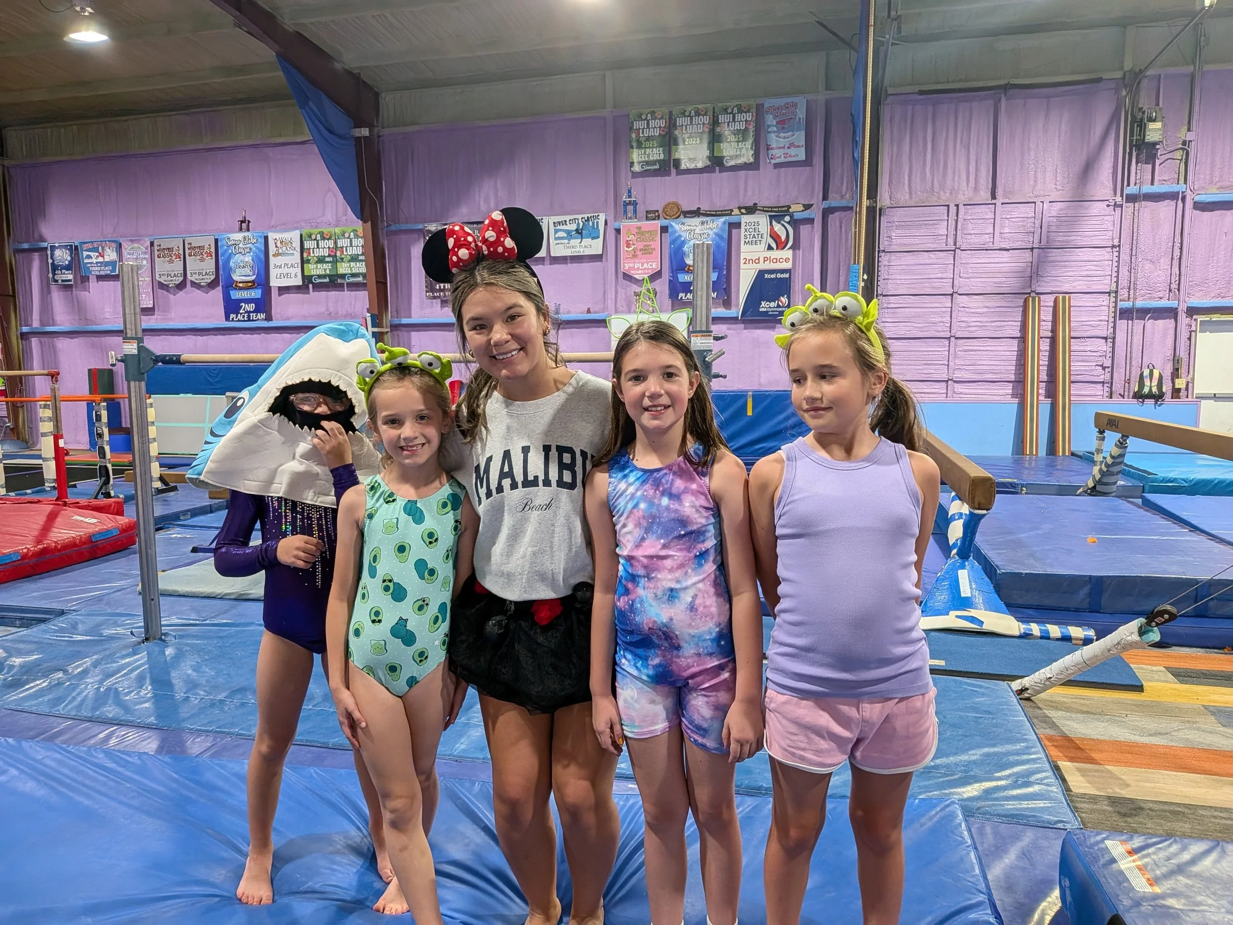 Five young girls and one woman posing inside a gymnastics gym, some of the girls are wearing costumes with alien and monster themes, one girl is dressed as a shark, and the woman is wearing Minnie Mouse ears. Hot Springs Gymnastics | 112 Bluestone Ct
