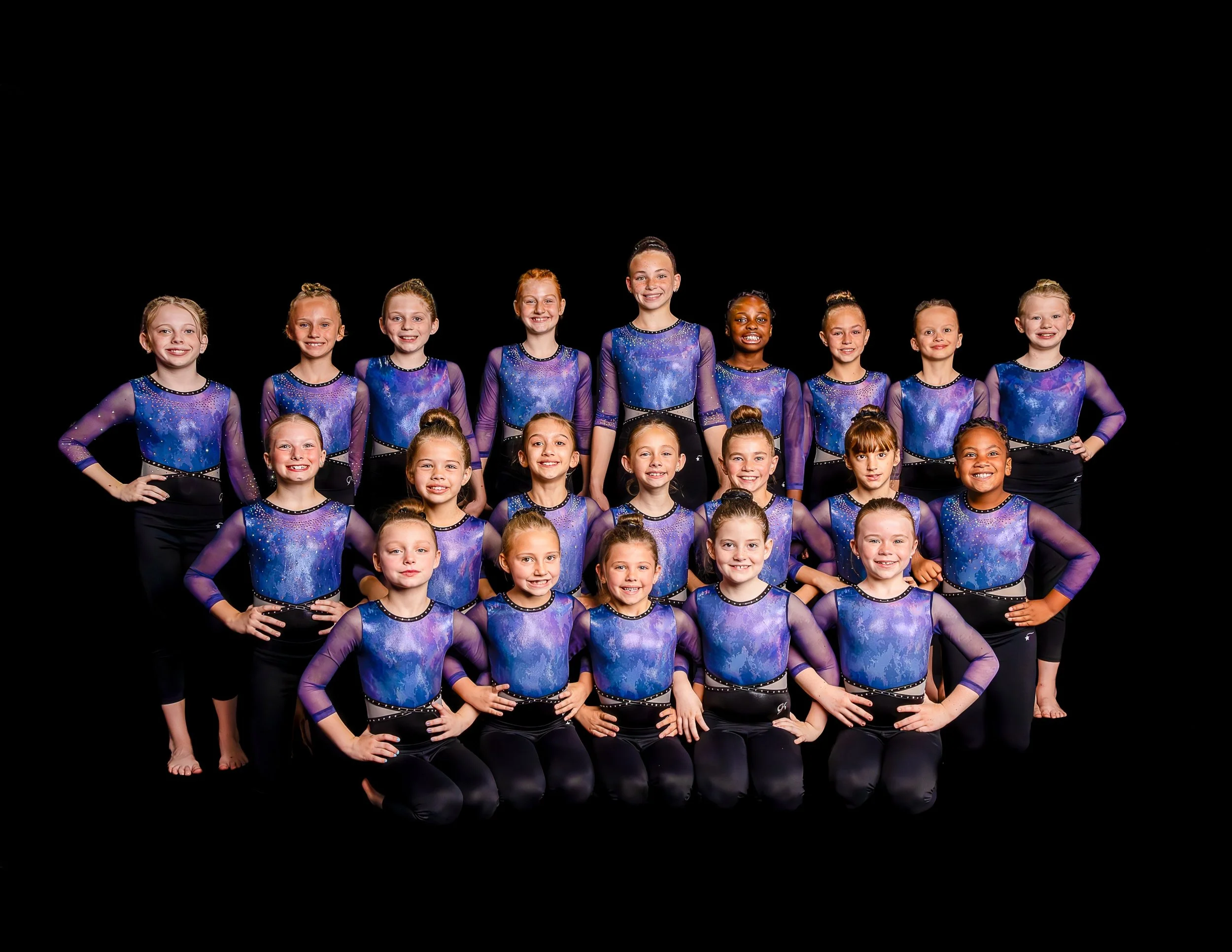 Group of young female gymnasts in blue and purple leotards standing together against black background.