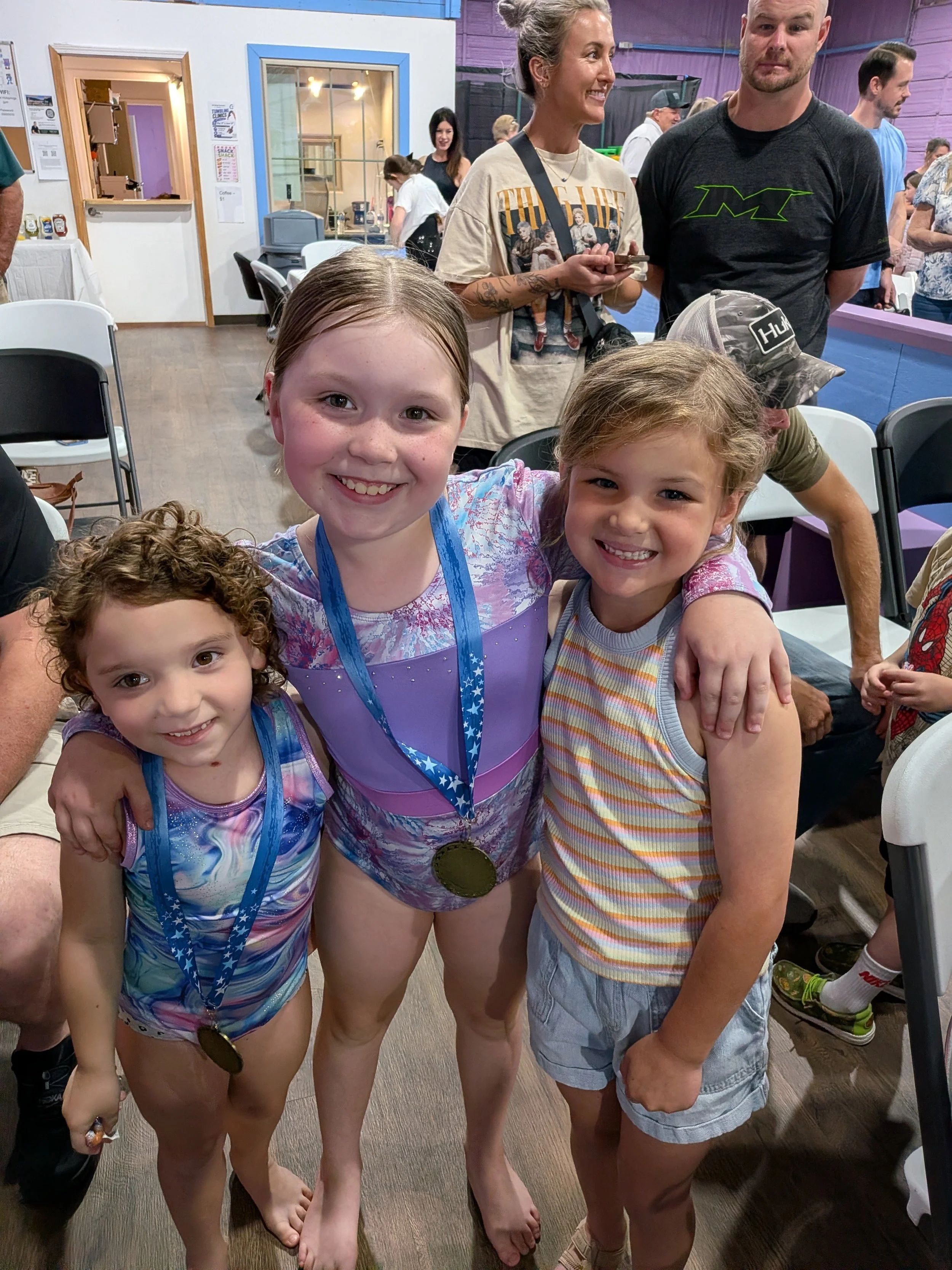 Three young girls standing together at an indoor event, smiling at the camera, wearing medals around their necks, and dressed in colorful athletic clothes. Behind them are adults and other children, with some seated at tables and others standing and chatting. The setting appears to be a recreational or community center.