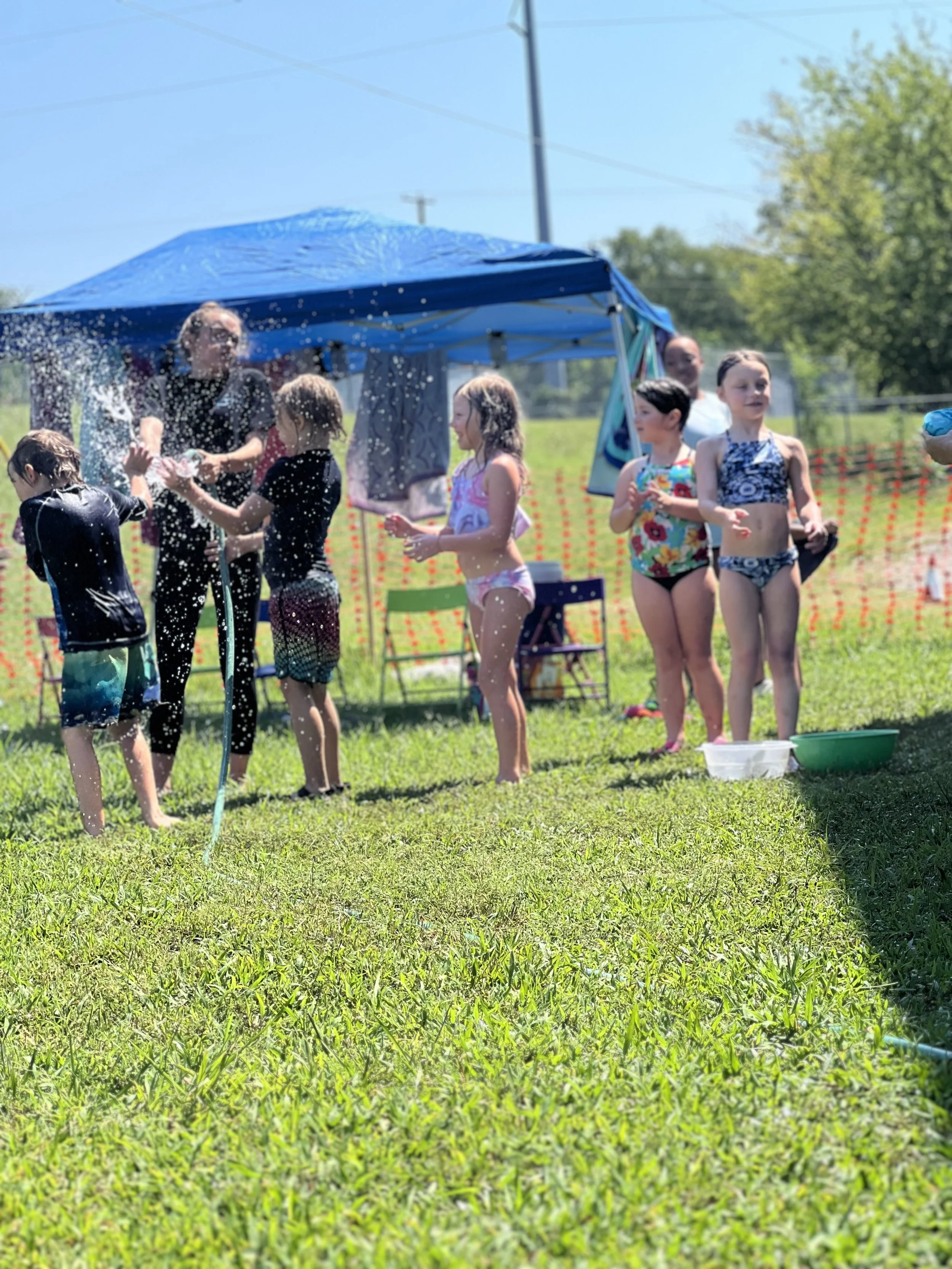 Children playing with water under a blue canopy on a grassy field on a sunny day.