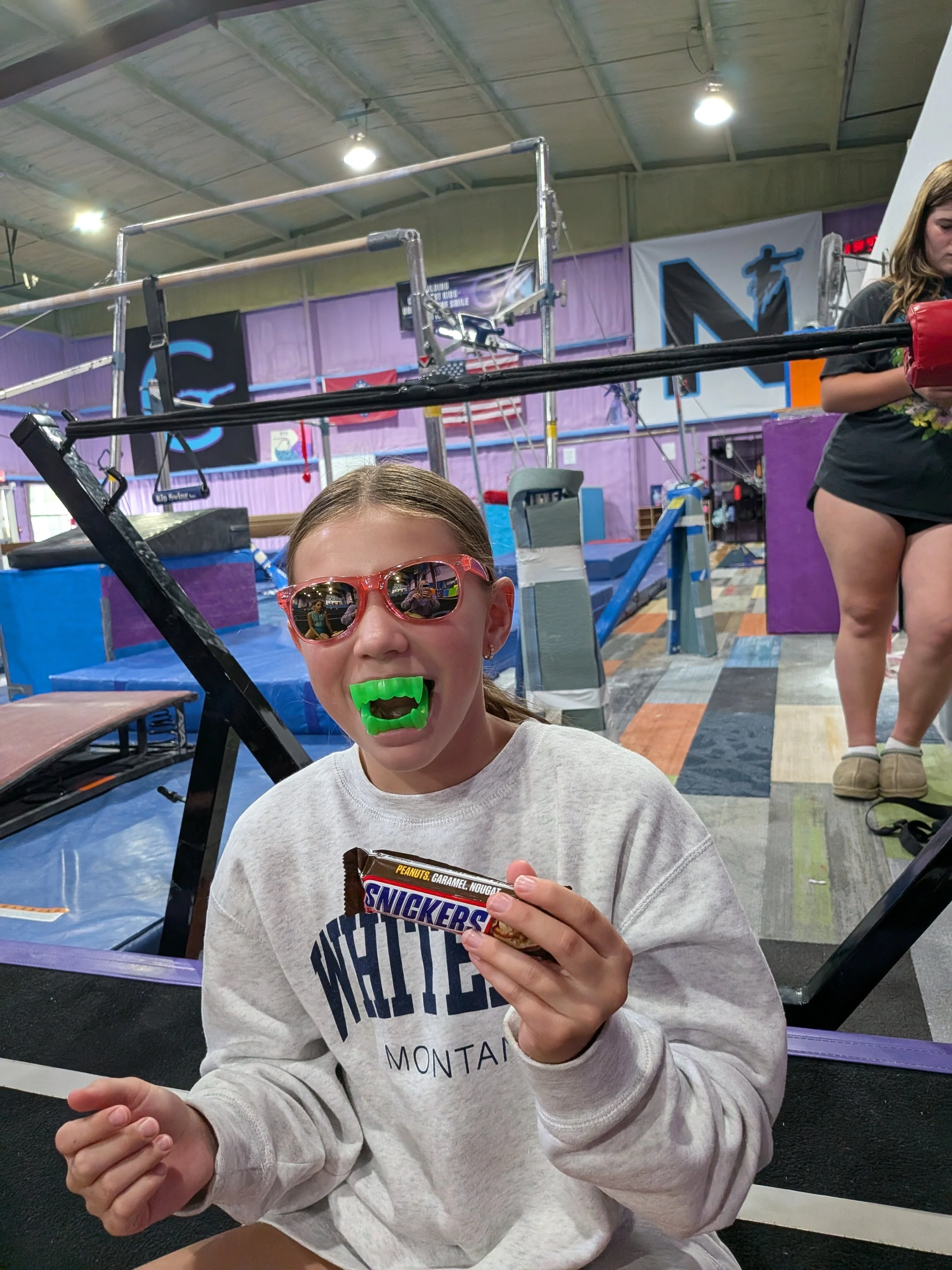 A young girl wearing pink sunglasses and a gray sweatshirt is holding a Snickers chocolate bar with a focus on her face and the chocolate in her hand inside an indoor gym or sports facility with various training equipment in the background.