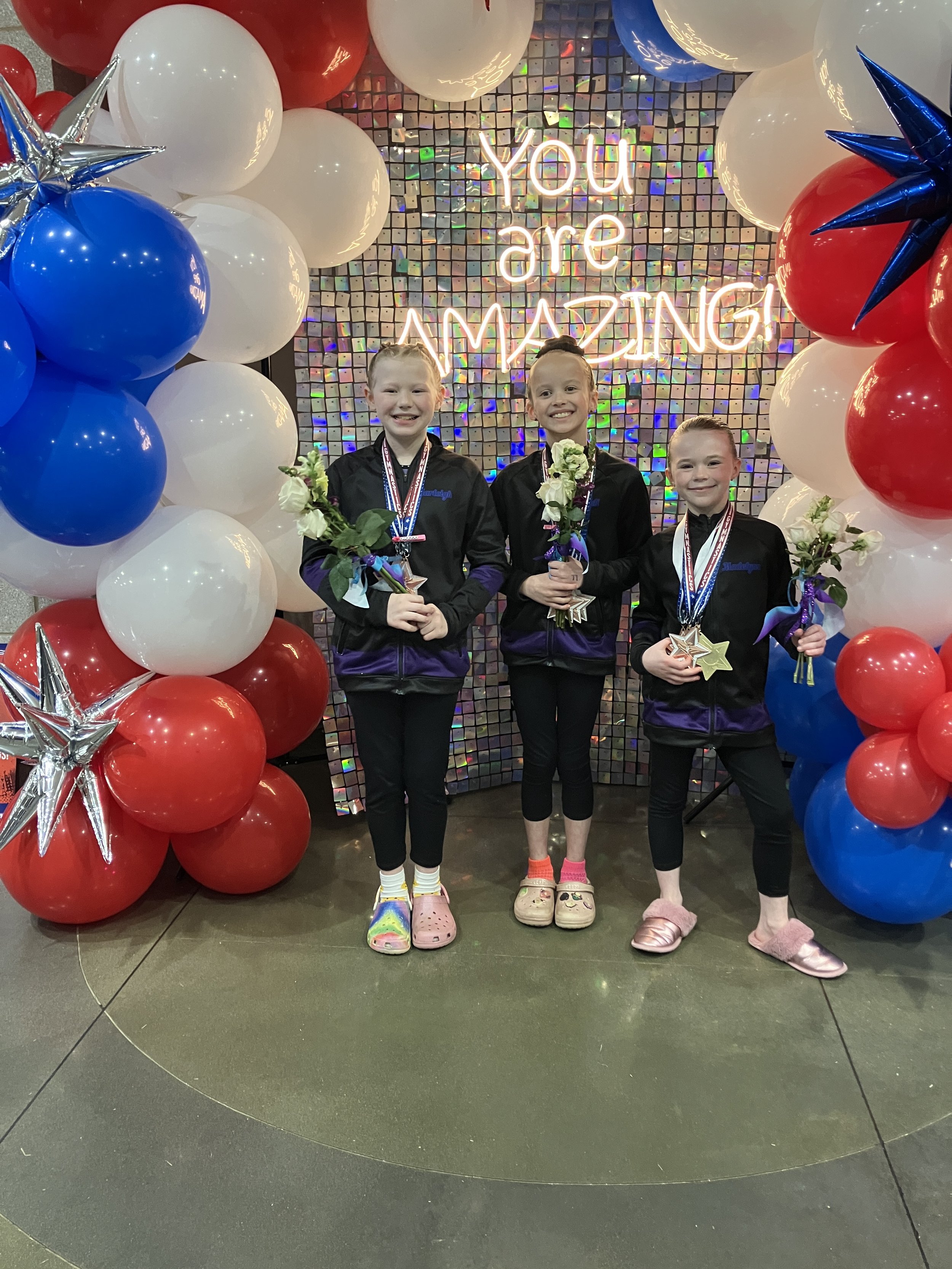 Three young girls wearing medals and holding flowers, standing in front of a colorful backdrop with a neon sign that says "You are AMAZING!" surrounded by red, white, and blue balloons and star-shaped balloons.