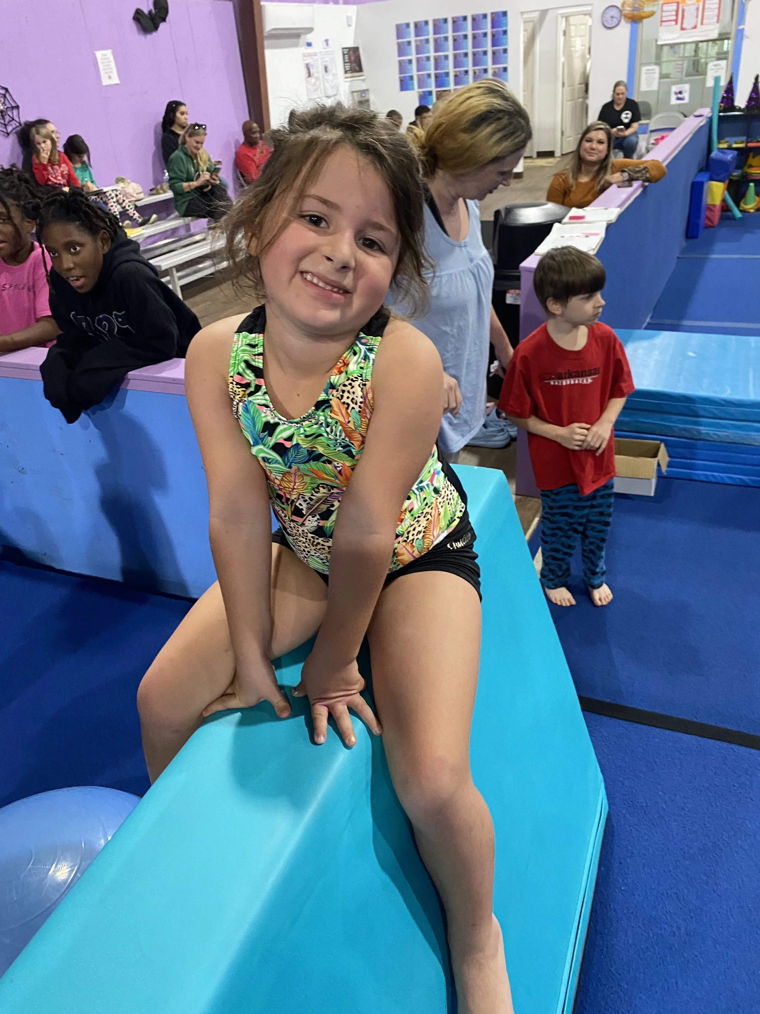 Young girl in colorful bathing suit sitting on a blue inflatable pool float at an indoor play area with children and adults in the background.