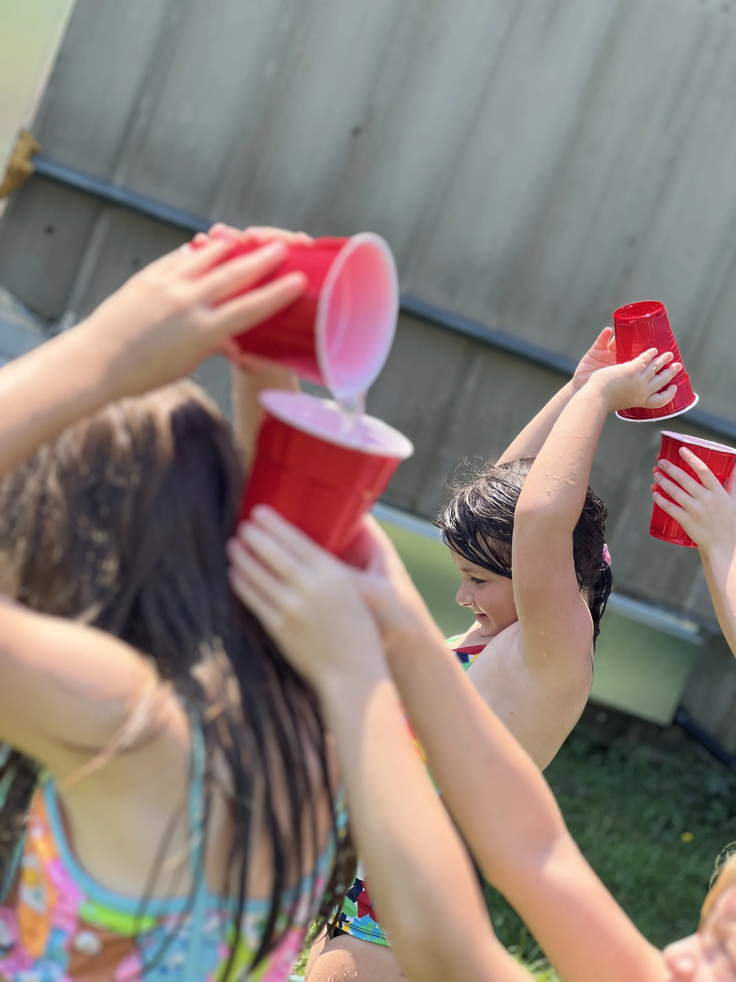 Children playing outdoors with red cups in a line, celebrating or participating in a water activity on a sunny day.
