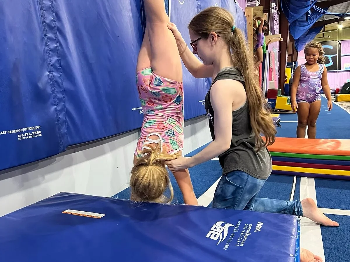 A girl in colorful shorts is upside down with her head on a padded mat and legs against a wall, while another girl kneels beside her, supporting her head. Two other girls, dressed in gymnastics attire, are in the background at a gymnastics gym.