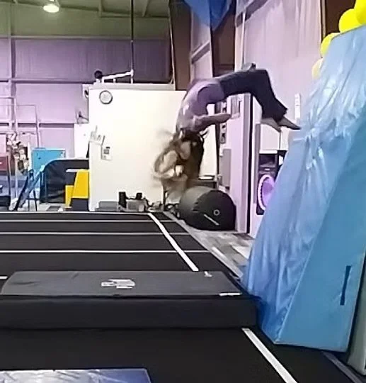 A person mid-air, performing a flip or acrobatic move in an indoor setting, with gym or industrial equipment in the background.  Hot Springs Gymnastics | 112 Bluestone Ct. Hot Springs, Arkansas 71901
