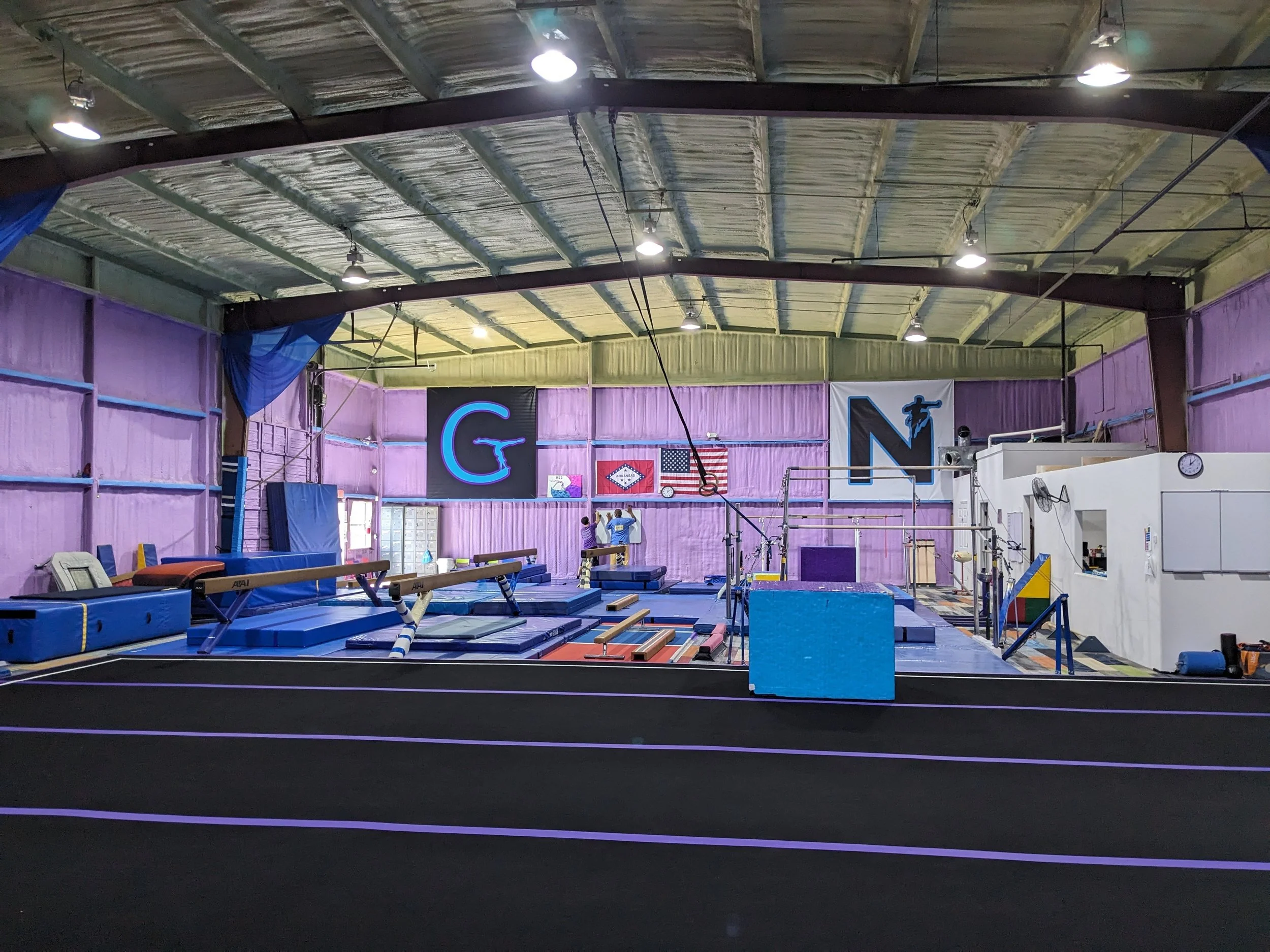Interior view of a gymnastics training facility with purple walls, black flooring with purple lines, and various gymnastics equipment including mats, bars, and foam blocks. Bright ceiling lights are visible, and there are two people practicing on the equipment.