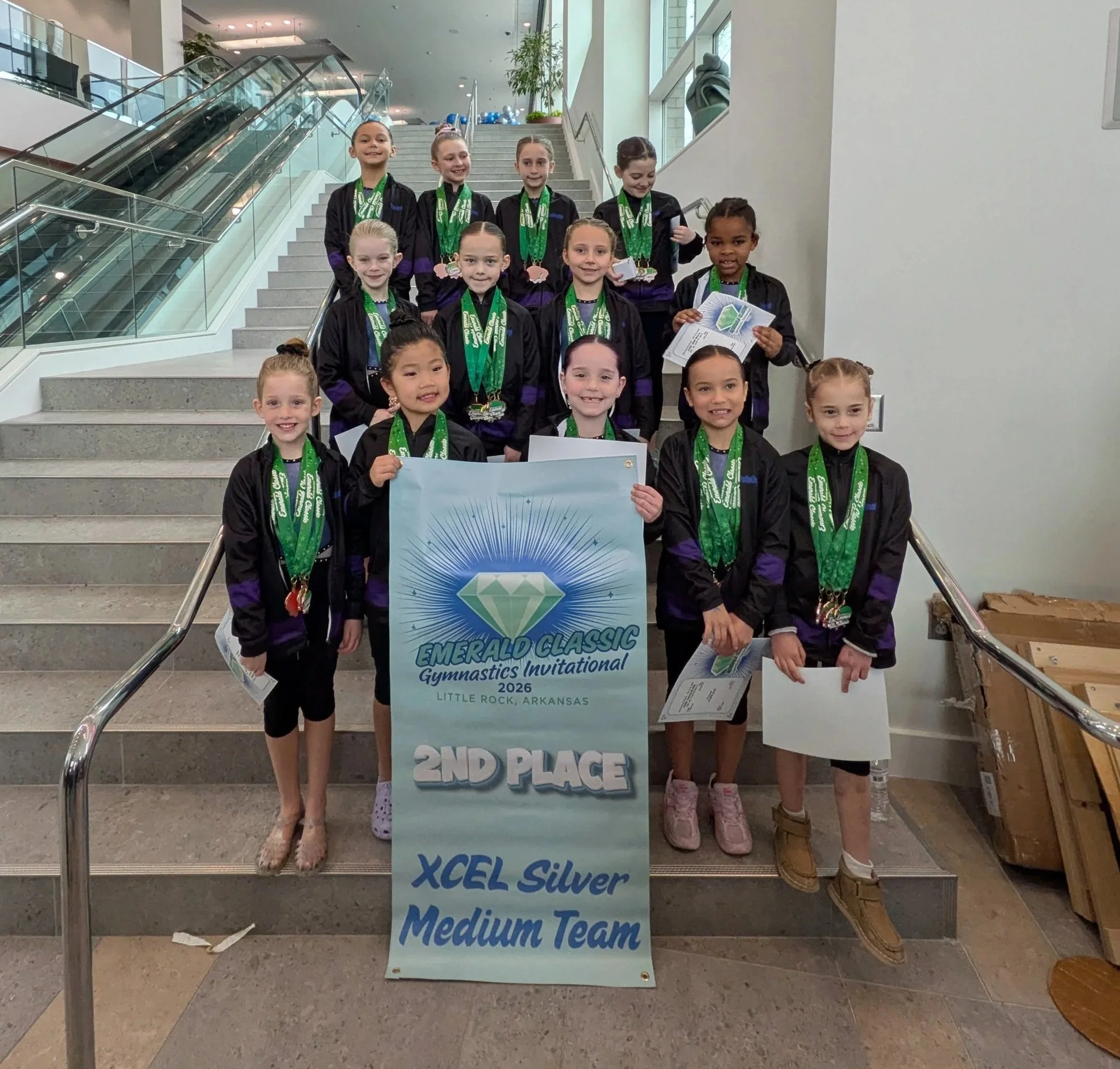 Group of young female gymnasts standing on a staircase holding a banner that reads 'Emerald Classic Gymnastics Invitational 2026 Little Rock Arkansas.' They are wearing black and purple jackets with medals around their necks, celebrating their second-place finish.