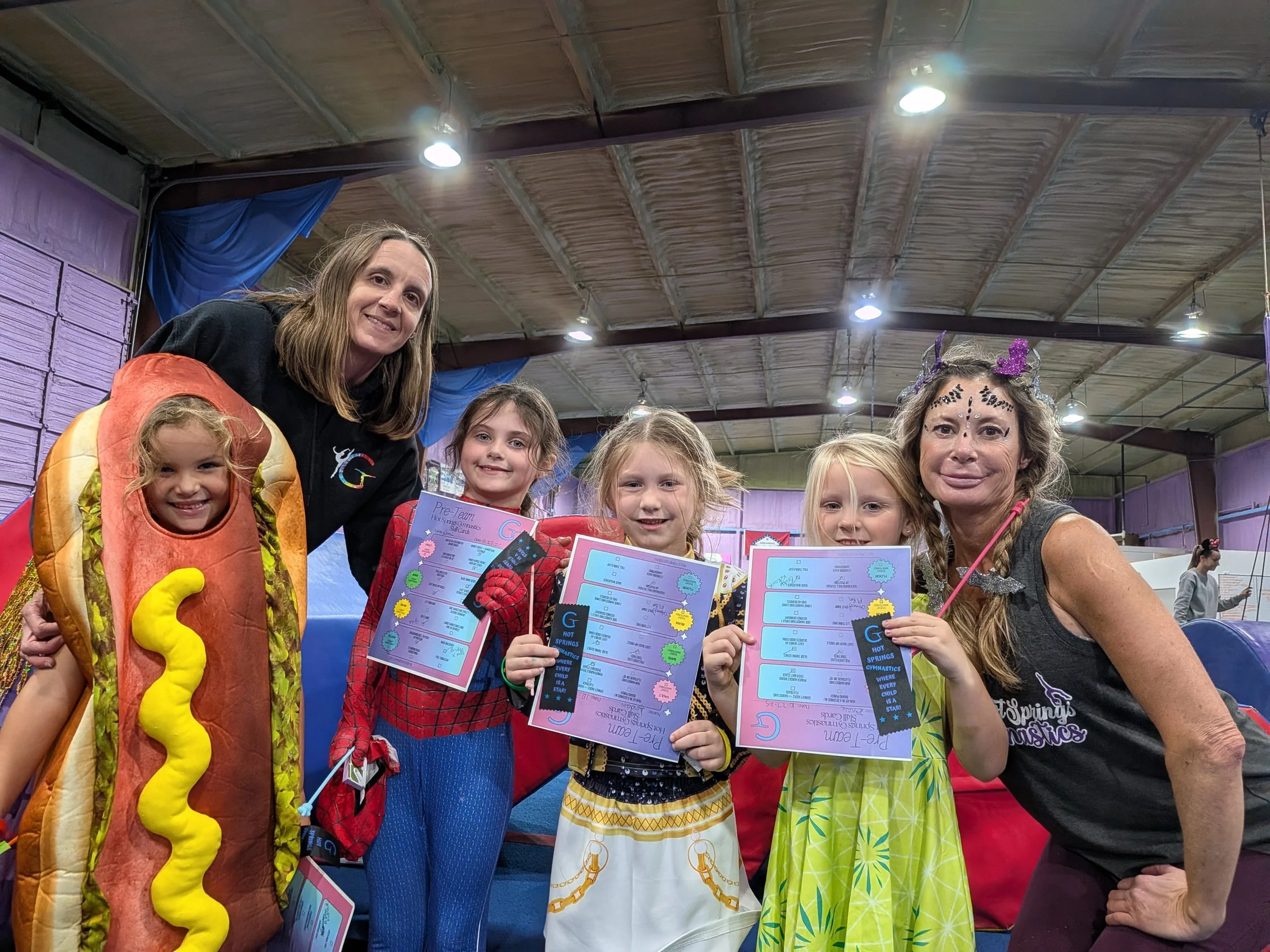 Group of children and two women at an indoor event, some children in costume including a hot dog and a taco, holding certificates, in a gymnasium with purple walls and blue curtain.