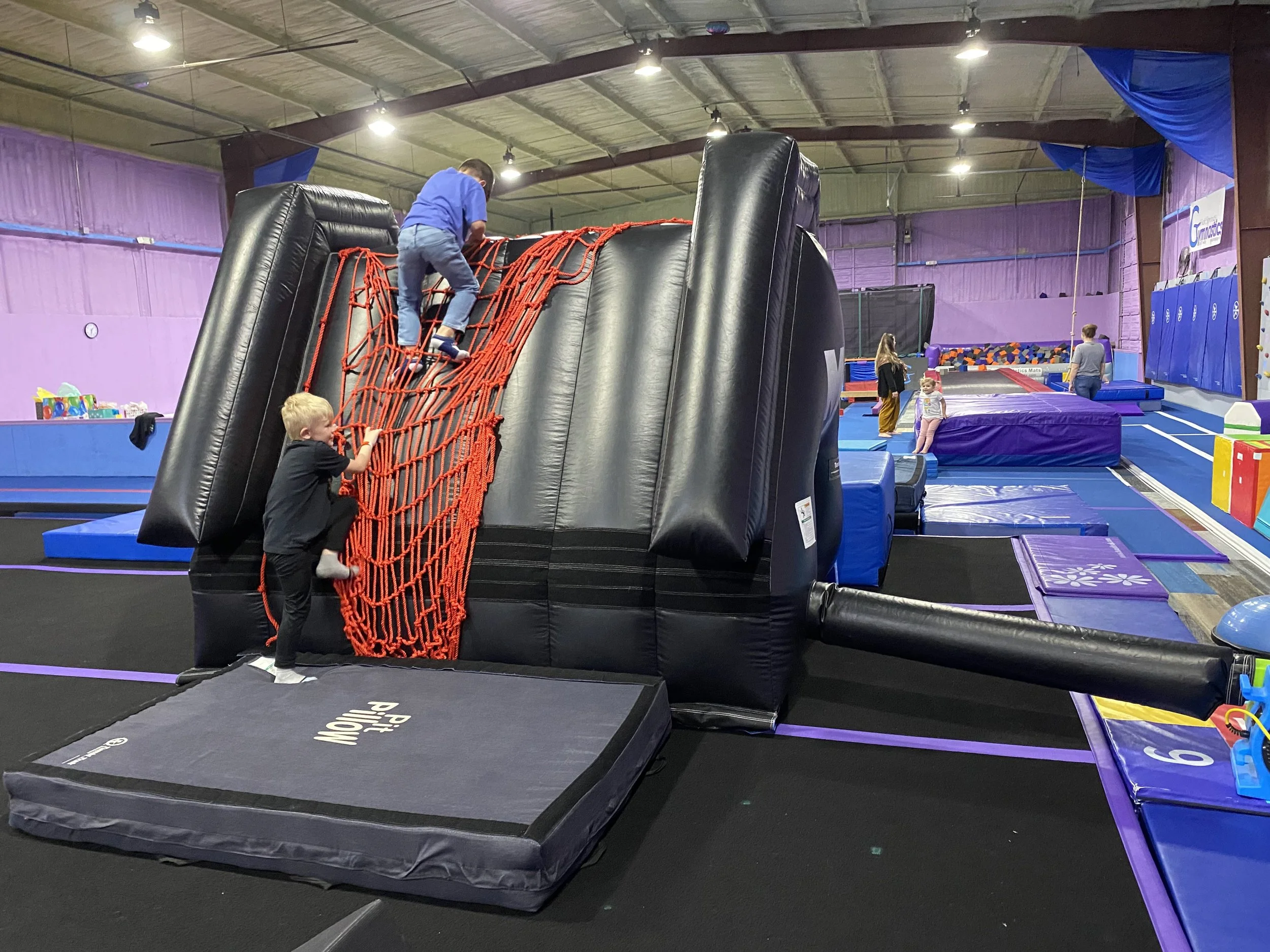 Children playing on an inflatable climbing wall inside an indoor trampoline park with purple walls and padded flooring.