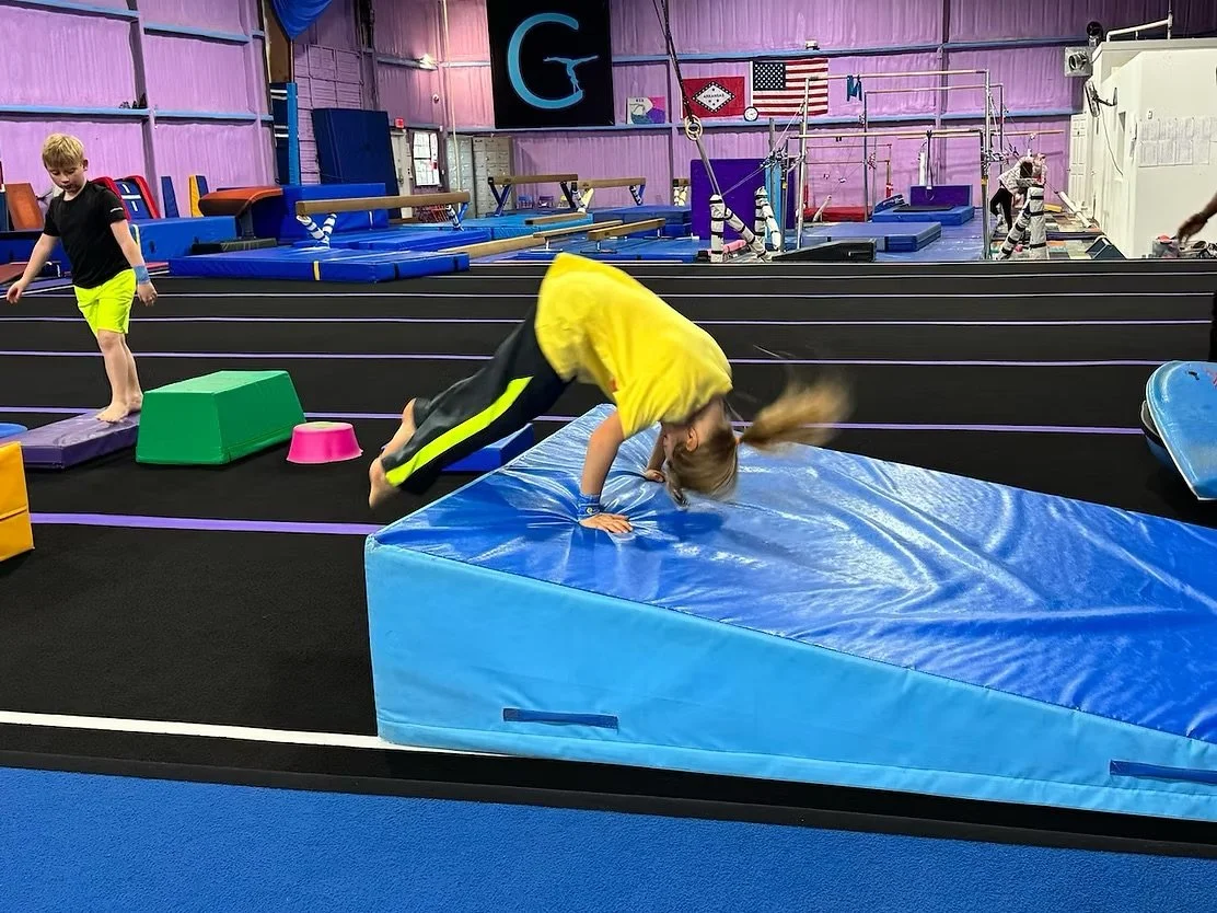 Kids practicing gymnastics in a training gym, with one girl in yellow clothes doing a forward flip over a blue padded mat, while another boy in black and yellow clothes stands nearby. Hot Springs Gymnastics | 112 Bluestone Ct. Hot Springs, Arkansas 7