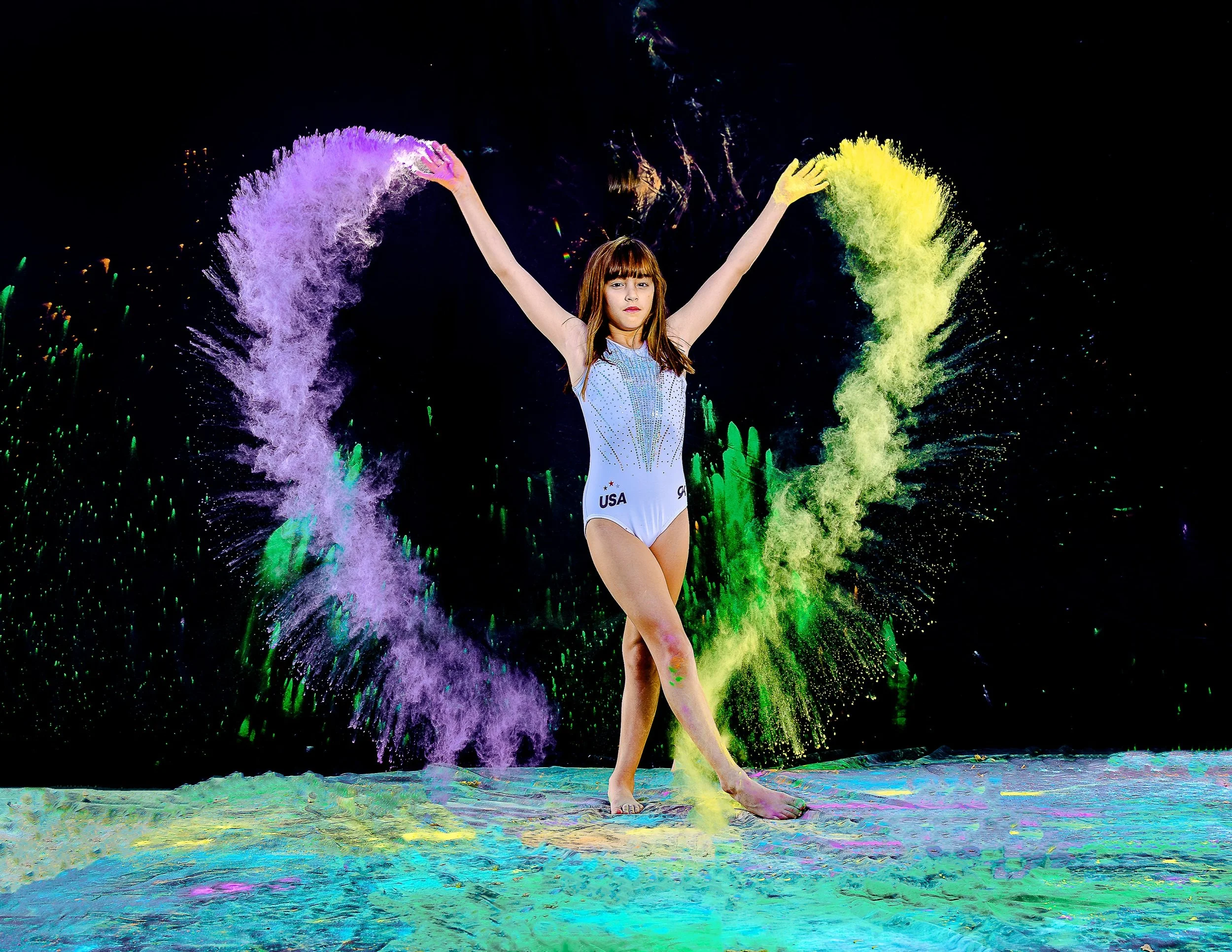 A female gymnast with the USA team uniform performing a handstand on a colorful floor, surrounded by vibrant powder explosion effects in purple, yellow, and green against a dark background.
