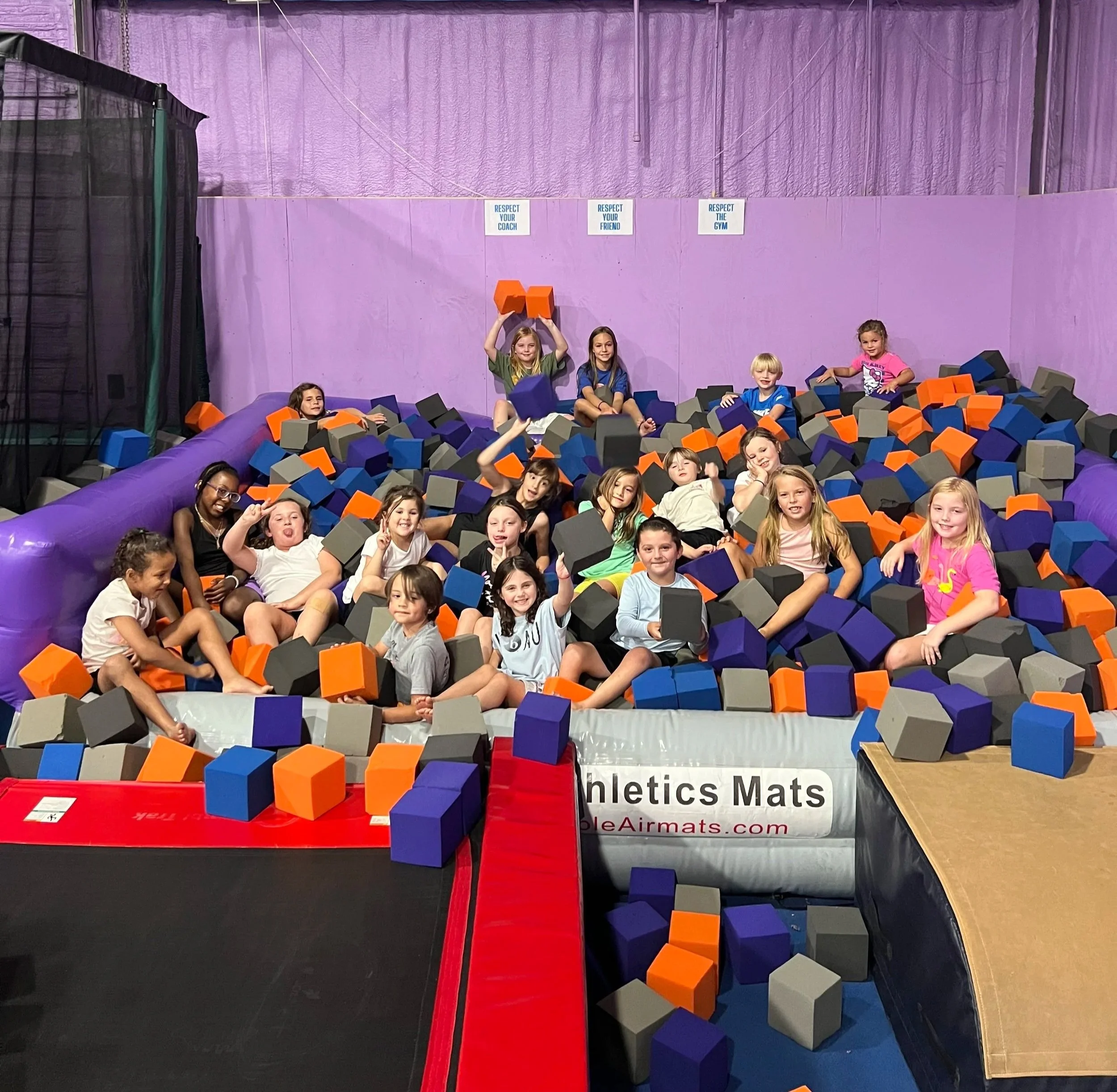 Children playing in an indoor foam pit with black, orange, gray, and purple foam blocks, purple walls, and signs on the wall that read respect your coach, respect your friend, and respect the gym.