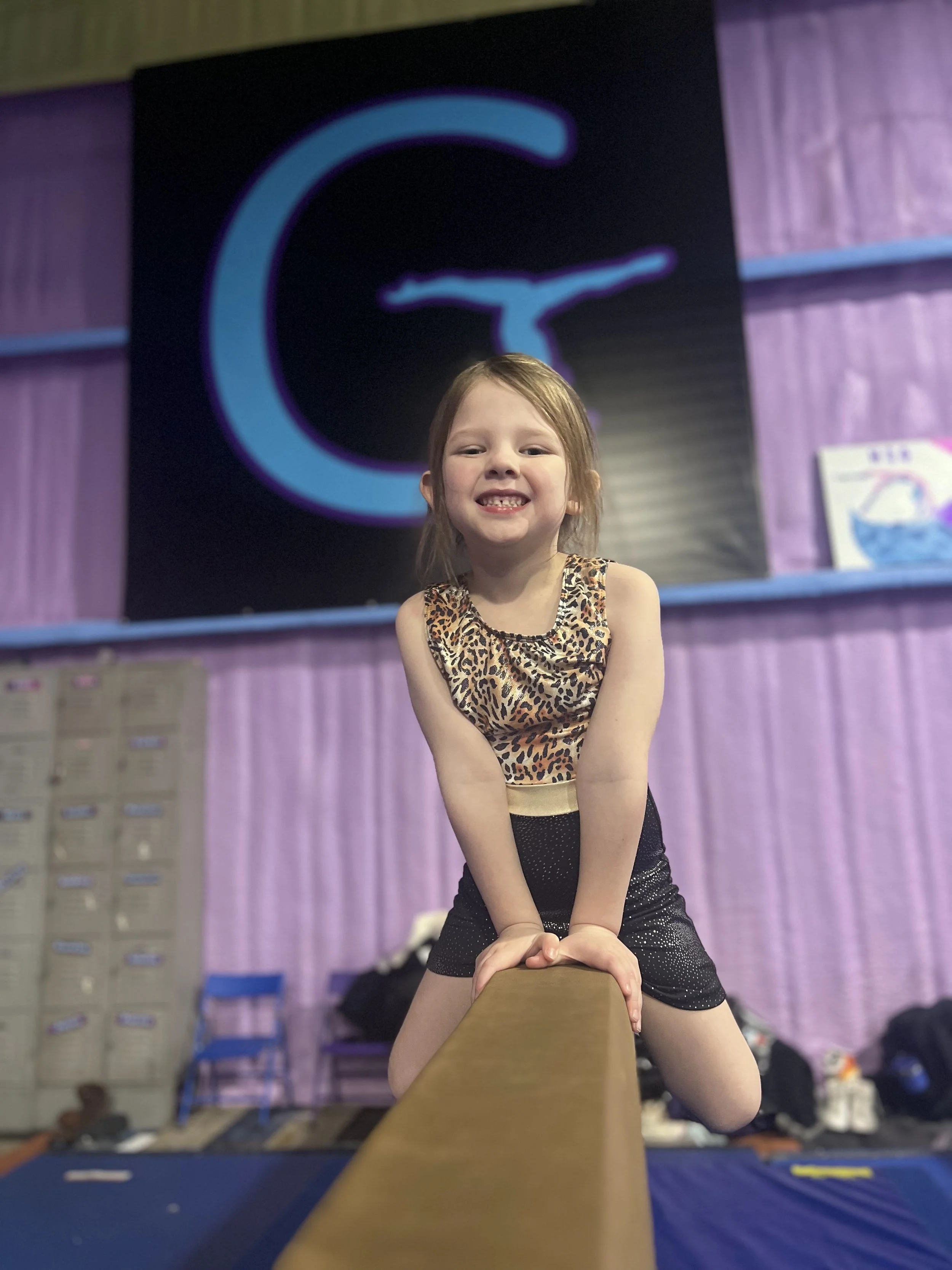 Young girl with long hair smiling, wearing a leopard-print top and shorts, performing a cheerleading stunt on a balance beam in a gymnasium with purple walls and a large logo in the background.